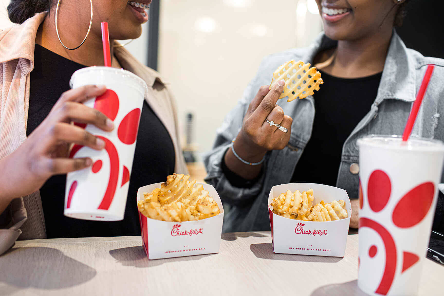 chick 桌布 壁紙 — 兩位微笑的女性在休閒餐桌上享用Chick-fil-A的格子薯條和飲料。