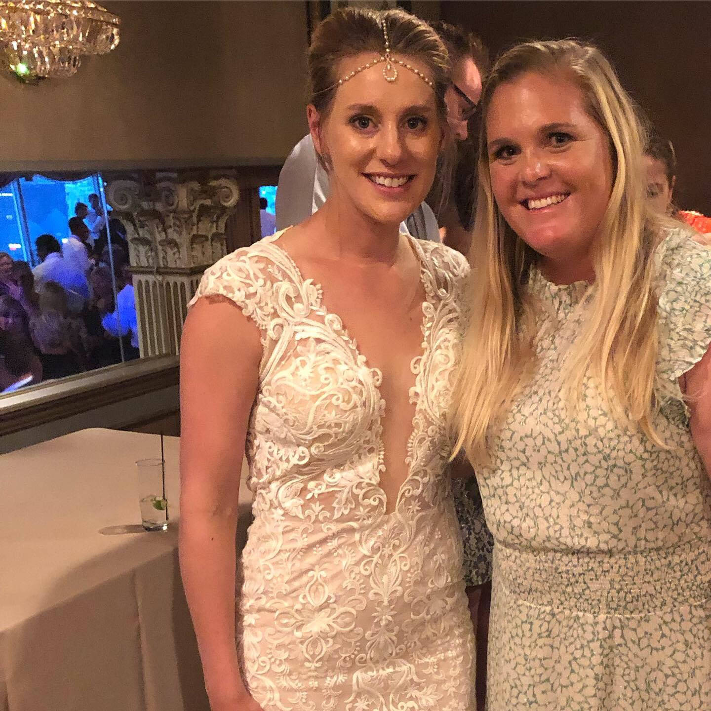 Wedding 桌布 壁紙 — 兩位微笑的女性在優雅的室內活動中一起擺姿勢，一位穿著精緻的白色蕾絲婚紗，另一位穿著花卉圖案連衣裙，上方有吊燈。