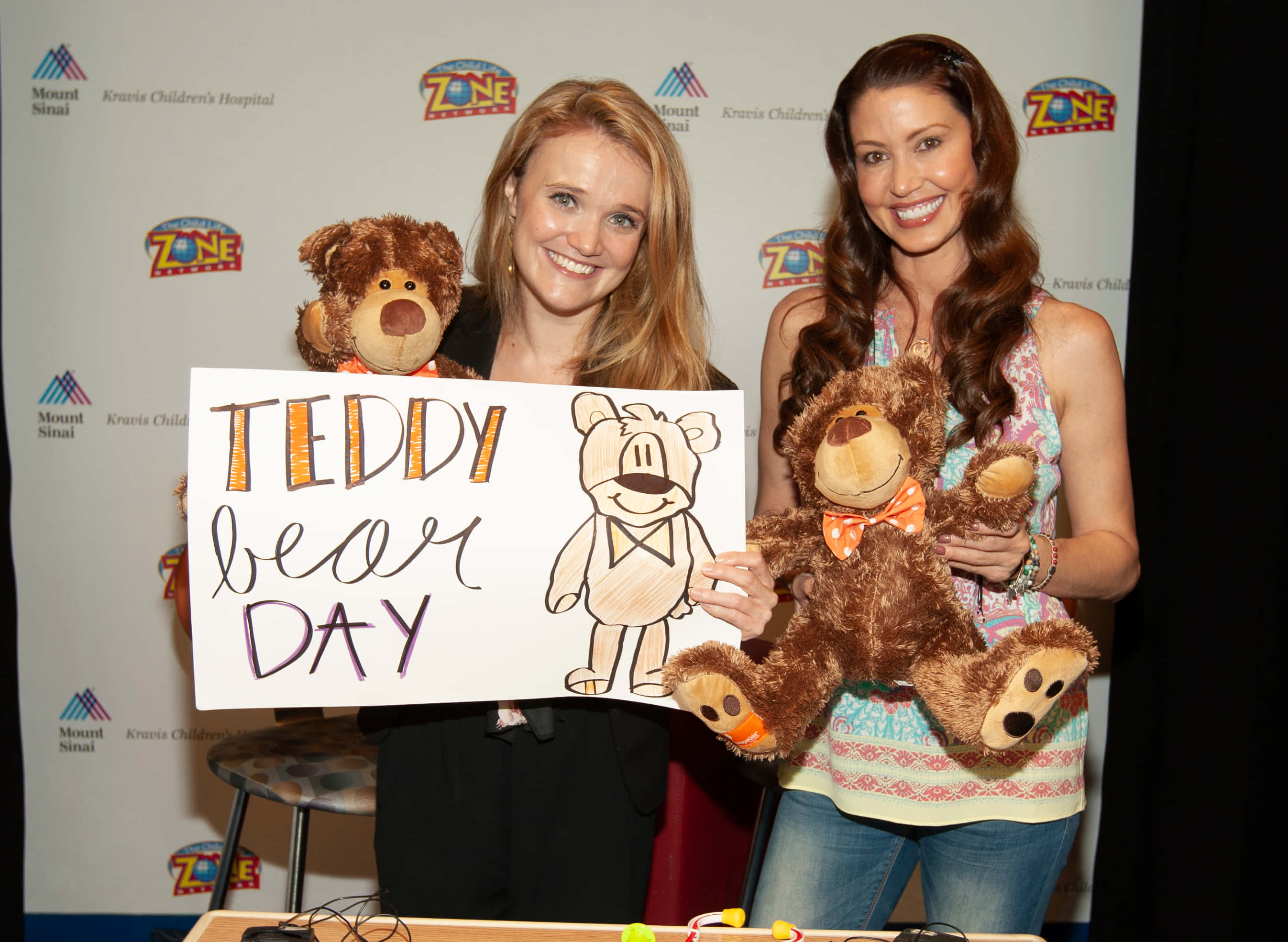 Shannon Elizabeth 桌布 壁紙 — 兩位微笑的女性在慈善活動中抱著泰迪熊和一個寫著「Teddy Bear Day」的標語牌。