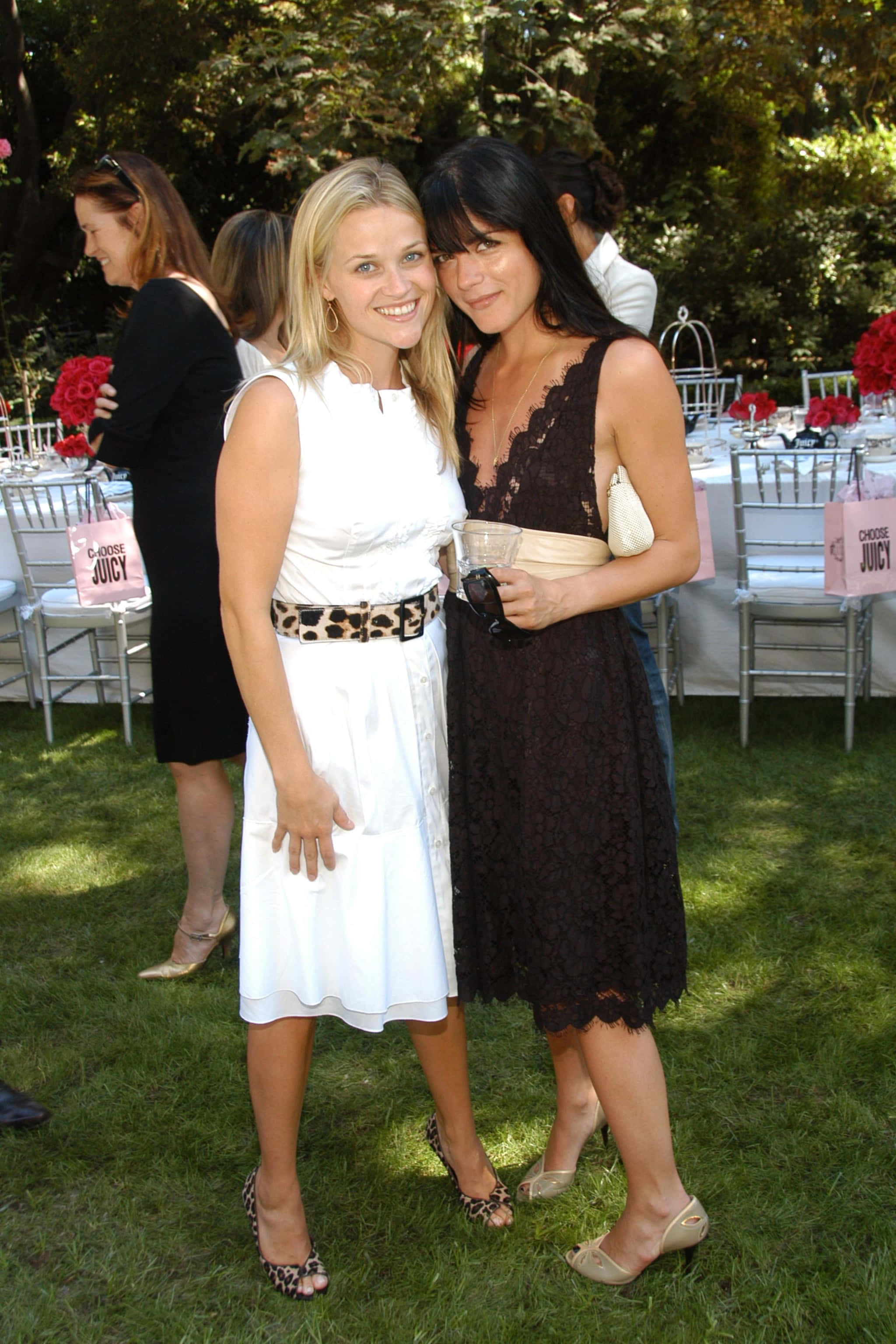 Selma Blair 桌布 壁紙 — 兩位微笑的女性在戶外花園派對的草地上一起擺姿勢，背景有桌子和紅花。