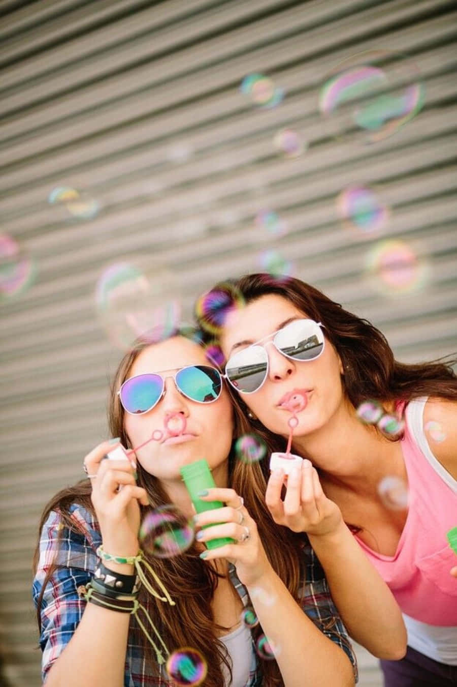 摯友 桌布 壁紙 — 兩位微笑的女性在波紋金屬背景前吹出色彩繽紛的肥皂泡，享受愉快的戶外時光。