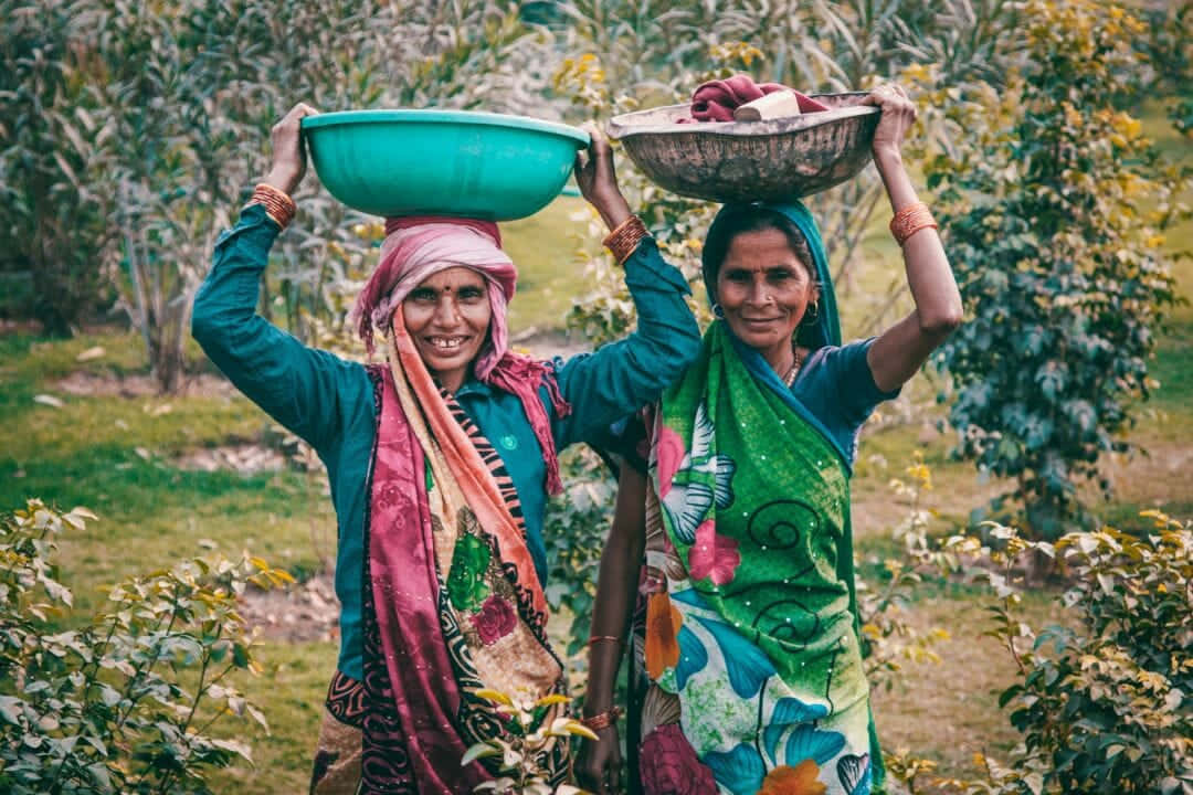 Indian 桌布 壁紙 — 兩位微笑的女性在鄉村環境中，在茂密的綠色樹葉間將彩色碗平衡在頭上。