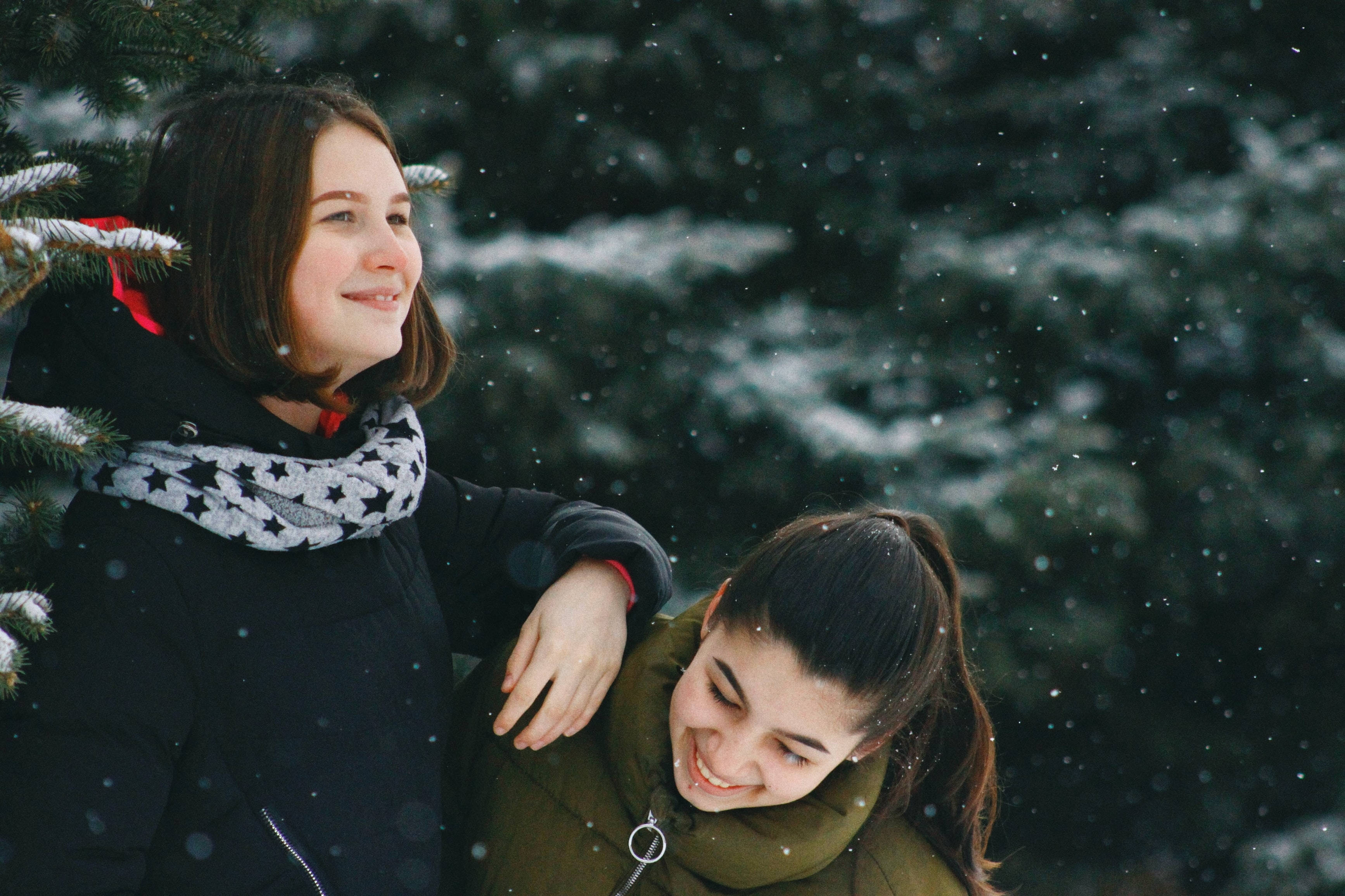 Teenage Girls 桌布 壁紙 — 兩位微笑的女性在飄雪中擁抱，旁邊是灑滿白雪的常青樹，身處寧靜的冬季森林。