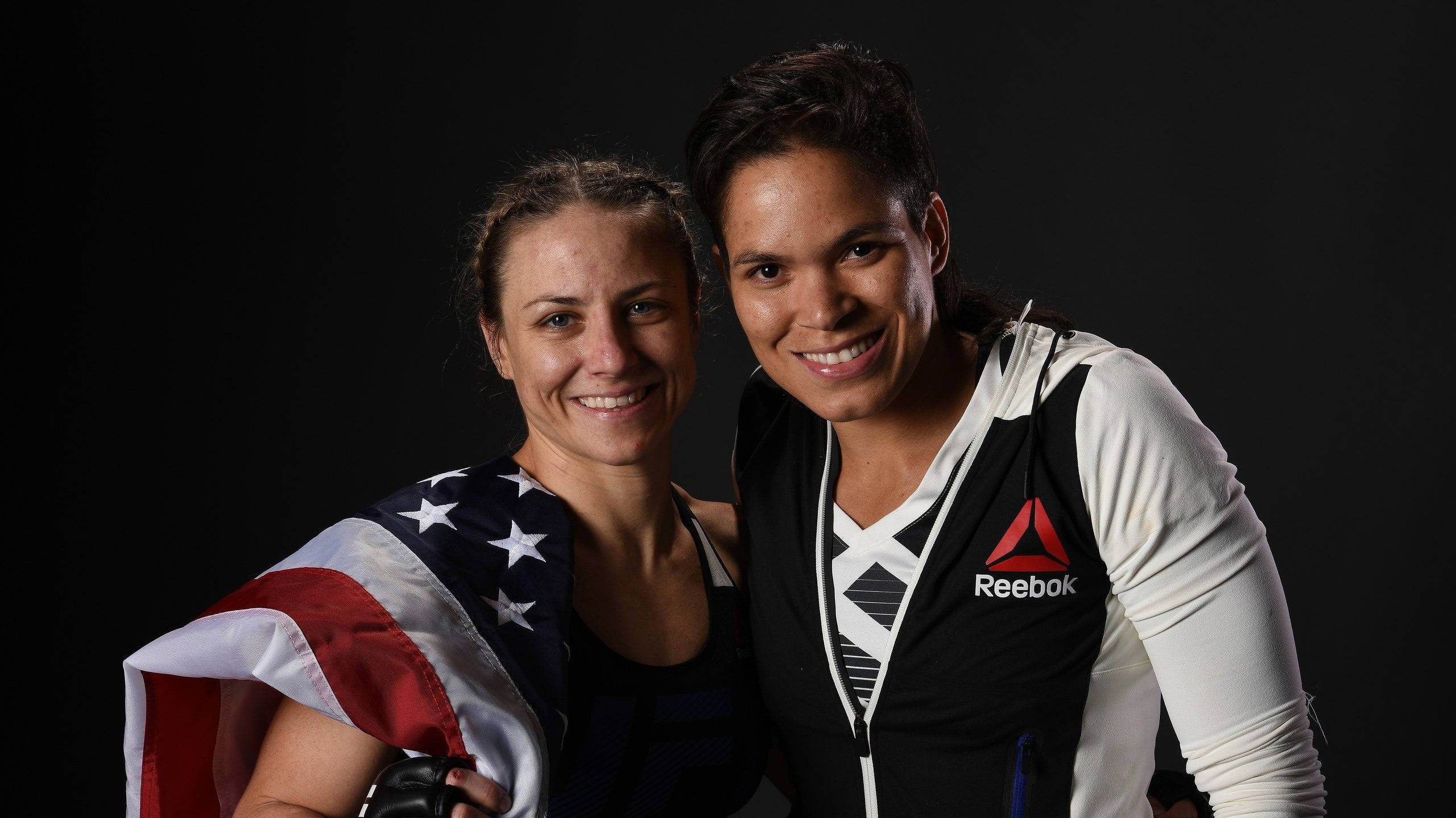MMA 桌布 壁紙 — 兩位微笑的女性拳手在黑色背景前緊密擺姿勢，一位披著美國國旗，另一位穿著Reebok運動背心。
