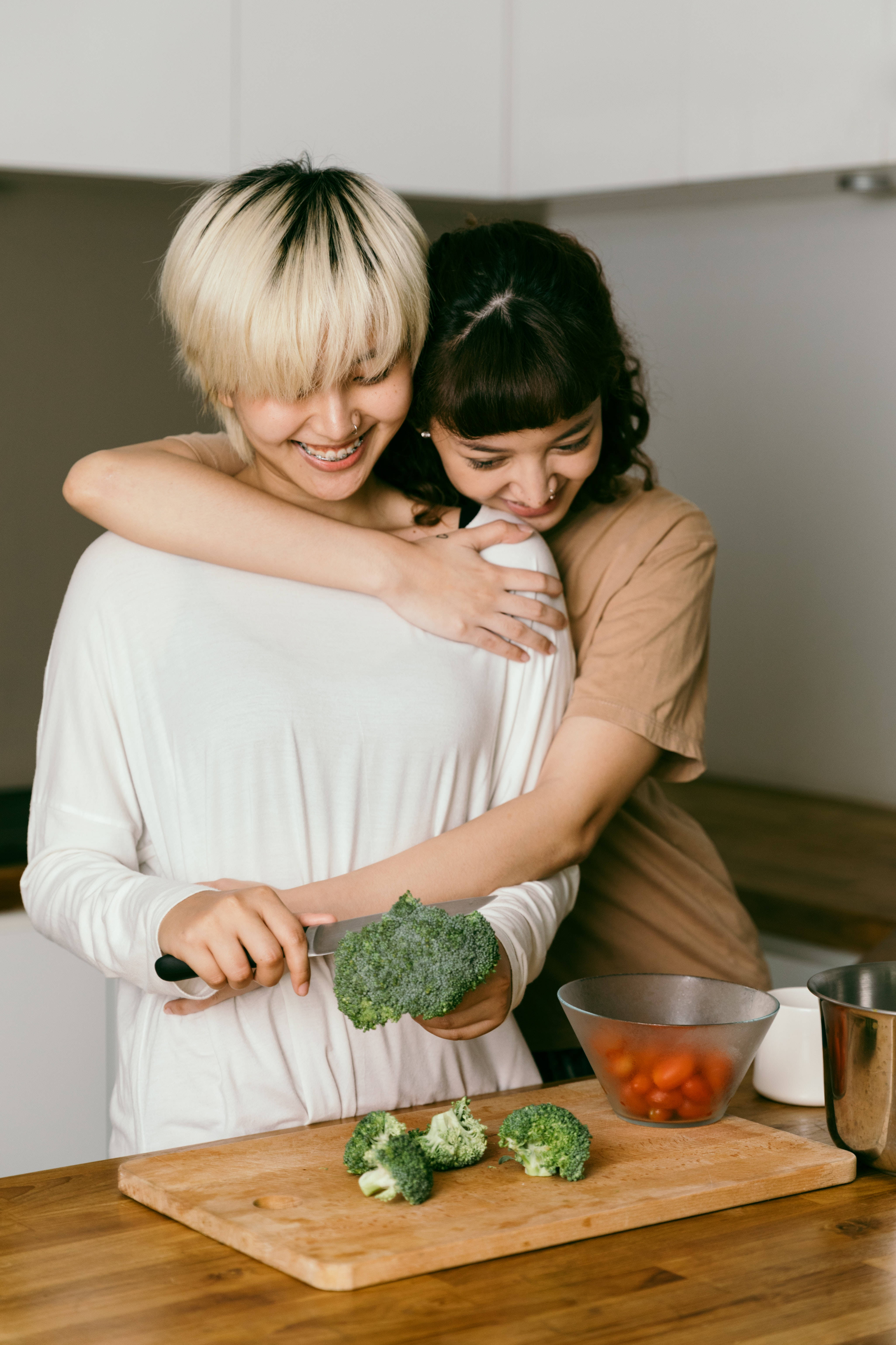 烹飪 桌布 壁紙 — 兩位微笑的女性熱情擁抱，其中一位在明亮的廚房的木製砧板上切花椰菜。