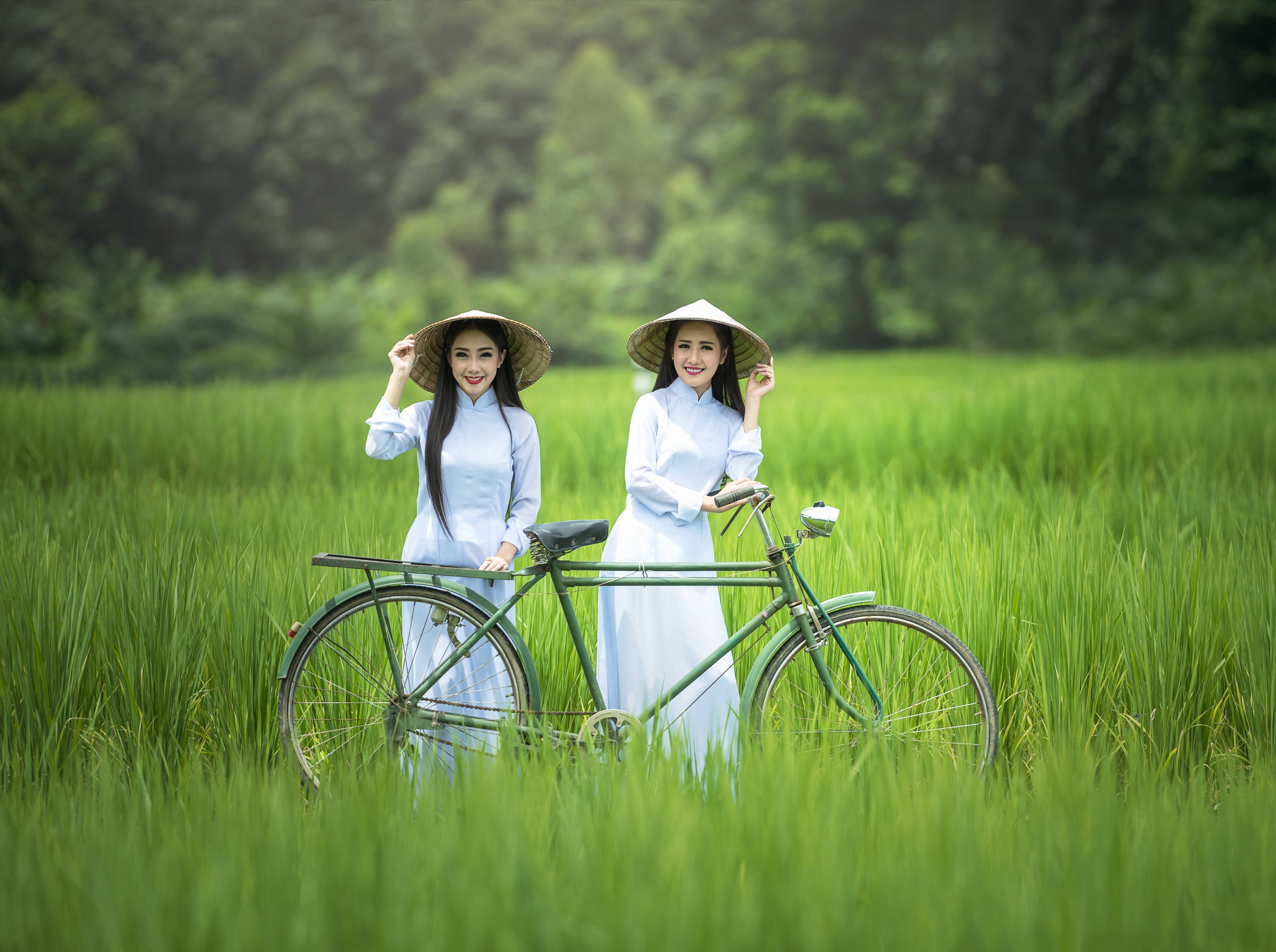Sister 桌布 壁紙 — 兩位微笑的女性穿著傳統白色奧黛和圓錐形帽子，站在一輛復古綠色自行車旁，背景是茂盛的綠色稻田。