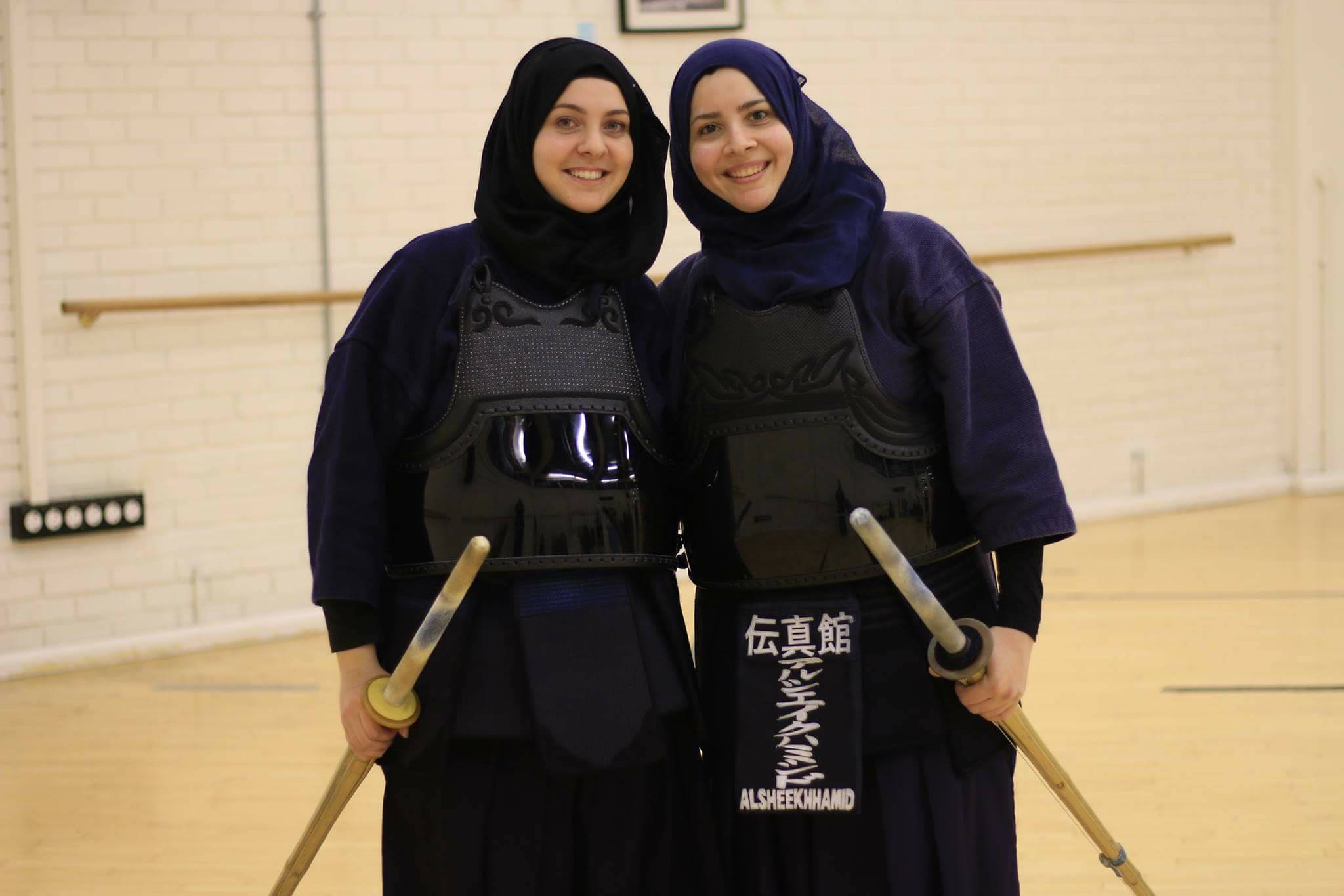 martial arts 桌布 壁紙 — 兩位微笑的女性穿著劍道裝備和頭巾，在體育館裡拿著竹劍擺姿勢。