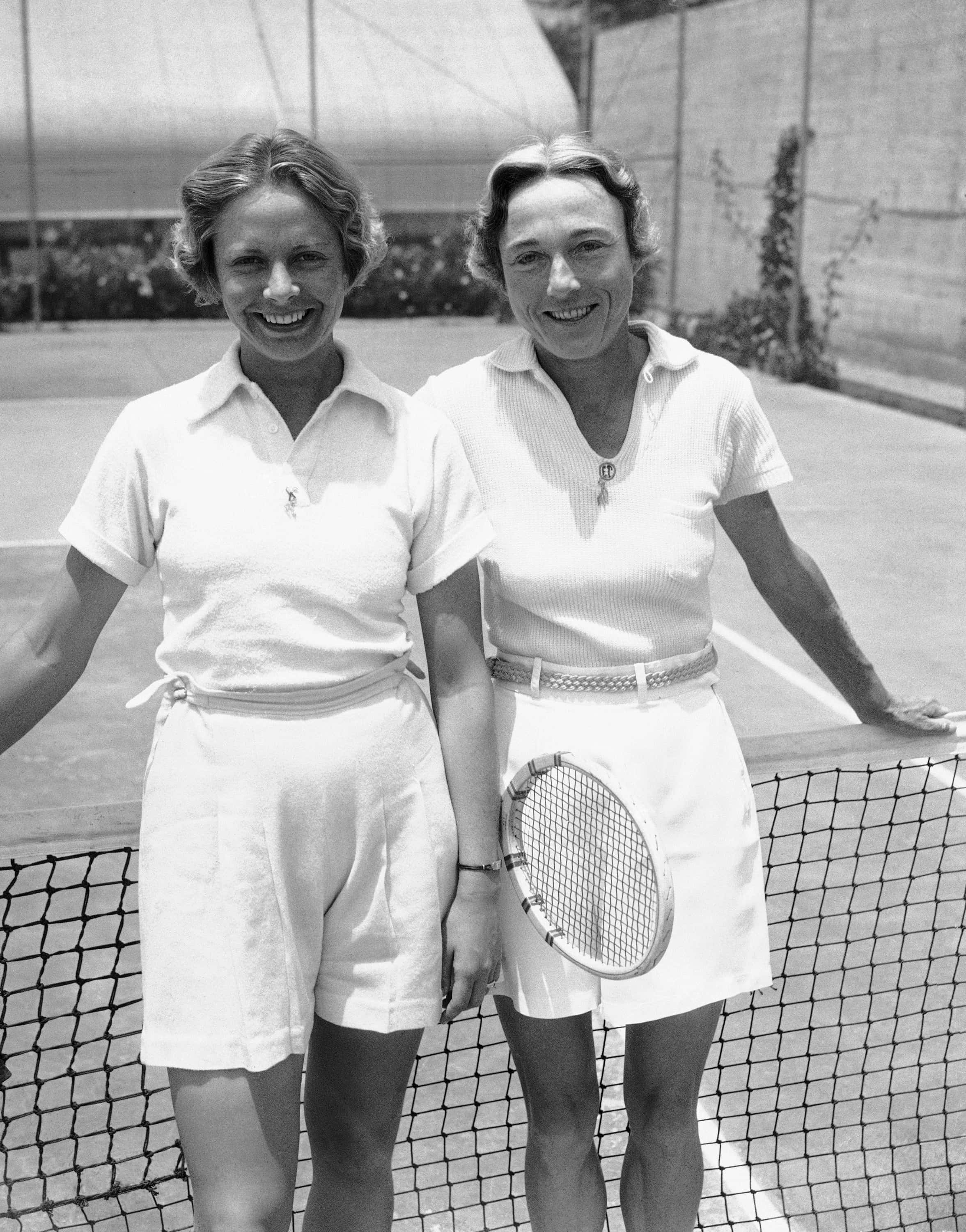 Tennis 桌布 壁紙 — 兩位微笑的女性穿著復古網球裝，在陽光明媚的戶外球場上，開心地站在網旁擺姿勢。