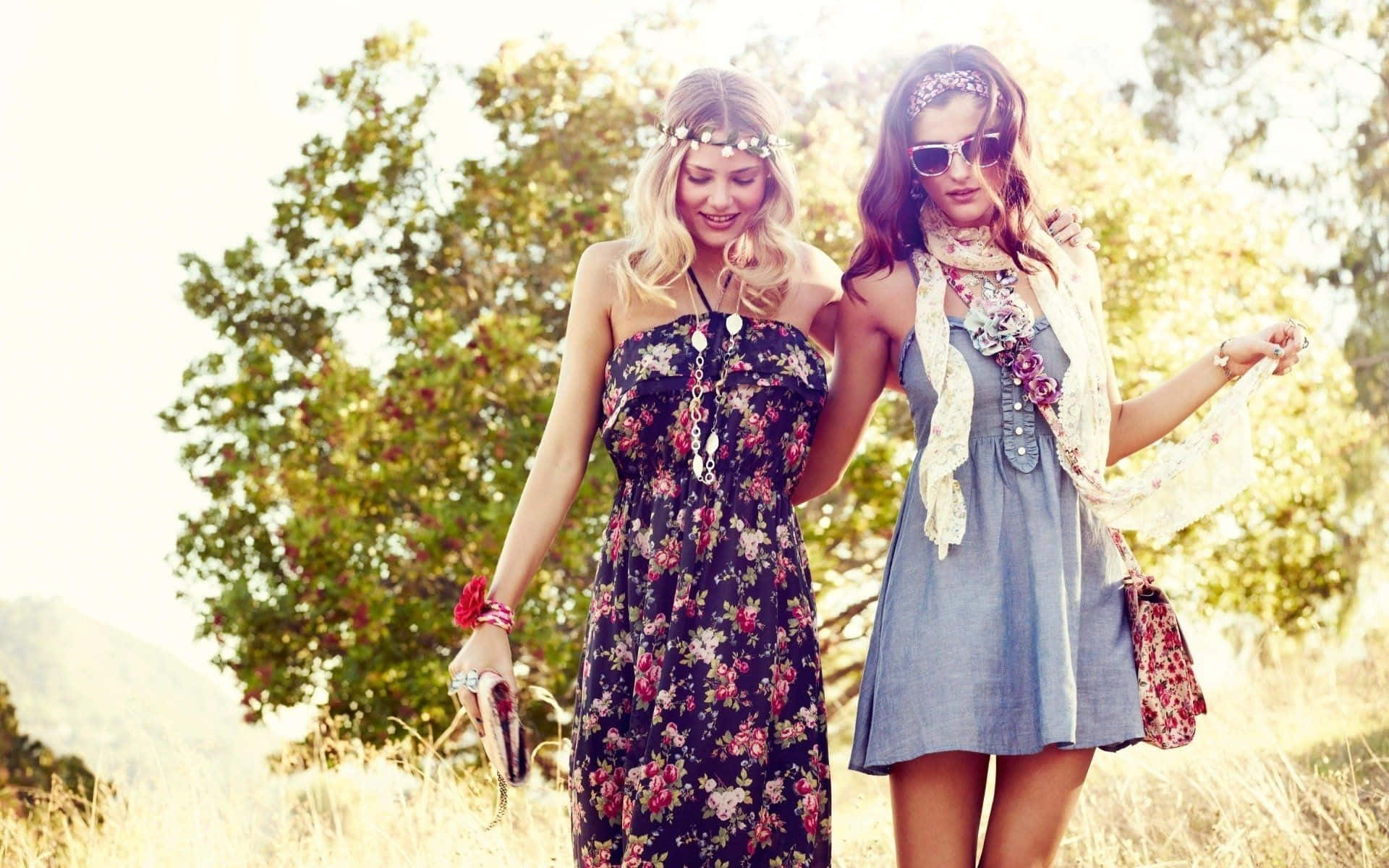 Spring Fashion 桌布 壁紙 — 兩位微笑的女性穿著波希米亞風格連衣裙，在陽光照耀的草地上漫步，身後有樹木和山丘。