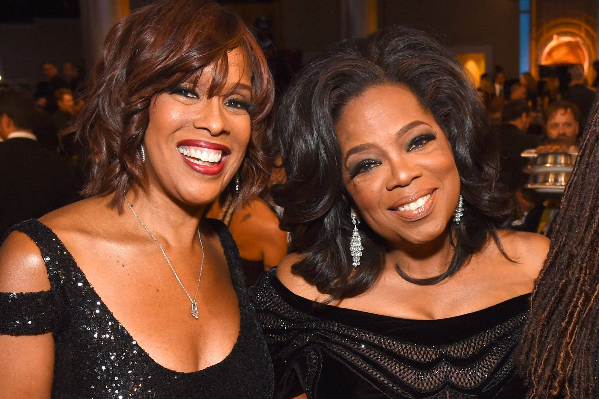 Oprah 桌布 壁紙 — 兩位微笑的女性穿著華麗的黑色晚禮服，在溫暖的環境燈光下，在擁擠的室內活動中一起擺姿勢。