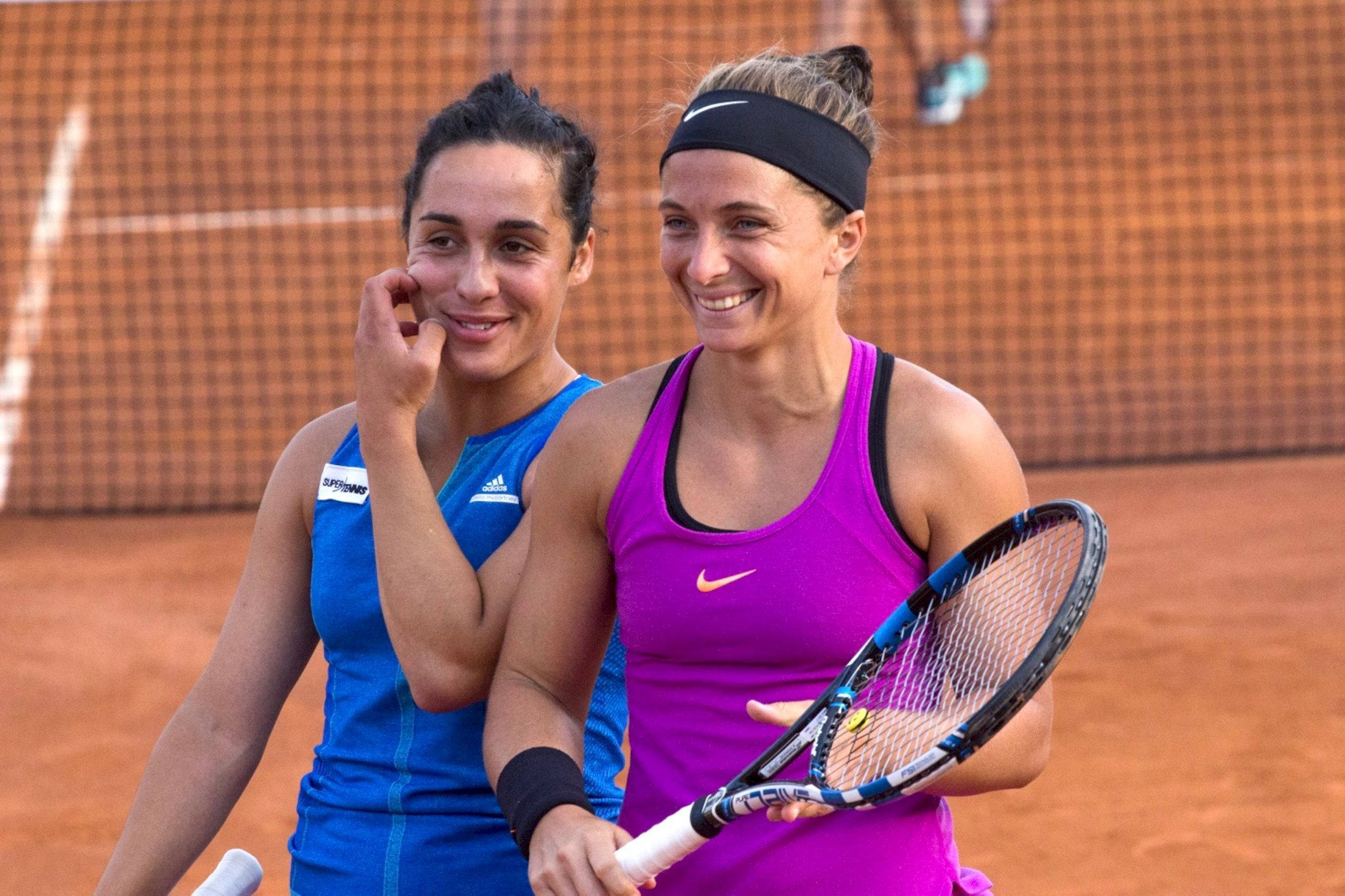 運動 桌布 壁紙 — 兩位微笑的女性網球選手在紅土球場上一起擺姿勢，一位穿藍色，一位穿紫色，手持球拍。