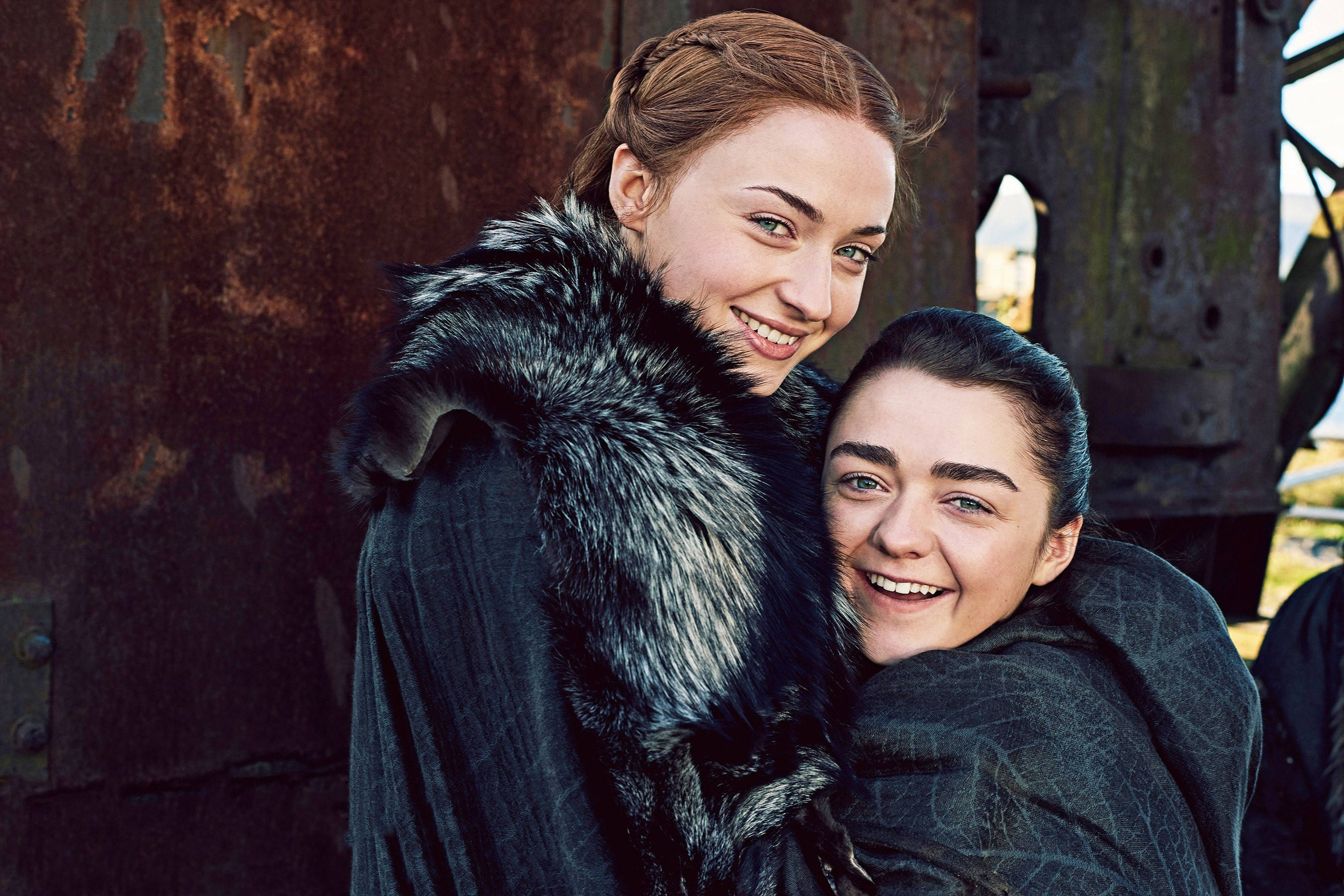 Sansa Stark 桌布 壁紙 — 兩位微笑的女性身穿帶毛皮的深色斗篷，在鏽蝕的金屬背景前溫暖地擁抱。