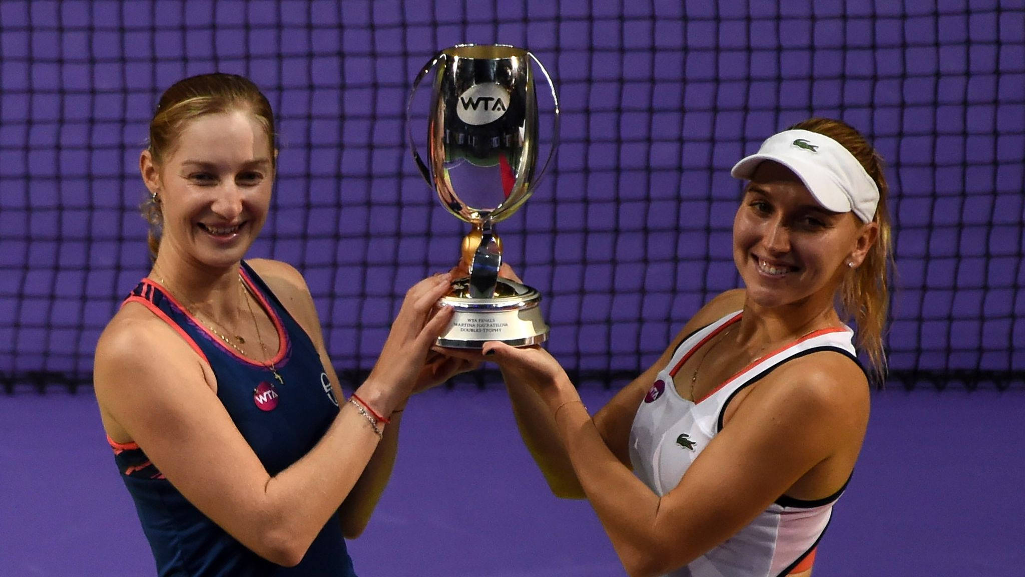 Tennis 桌布 壁紙 — 兩位微笑的女網球選手一起舉起閃亮的WTA獎盃，背景是紫色球場網。