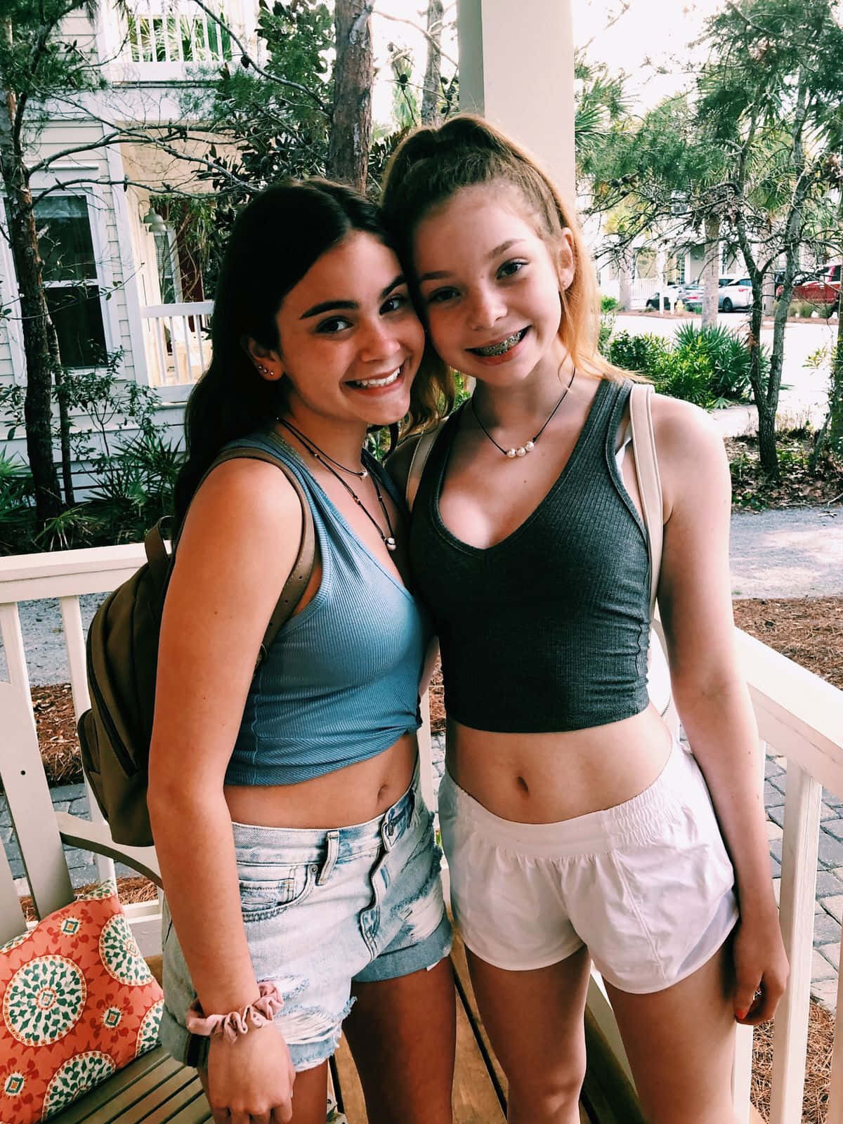 girls 桌布 壁紙 — 兩位微笑的年輕女性在明亮的日光下，於被綠植和白色房屋環繞的門廊上緊密地擺姿勢。