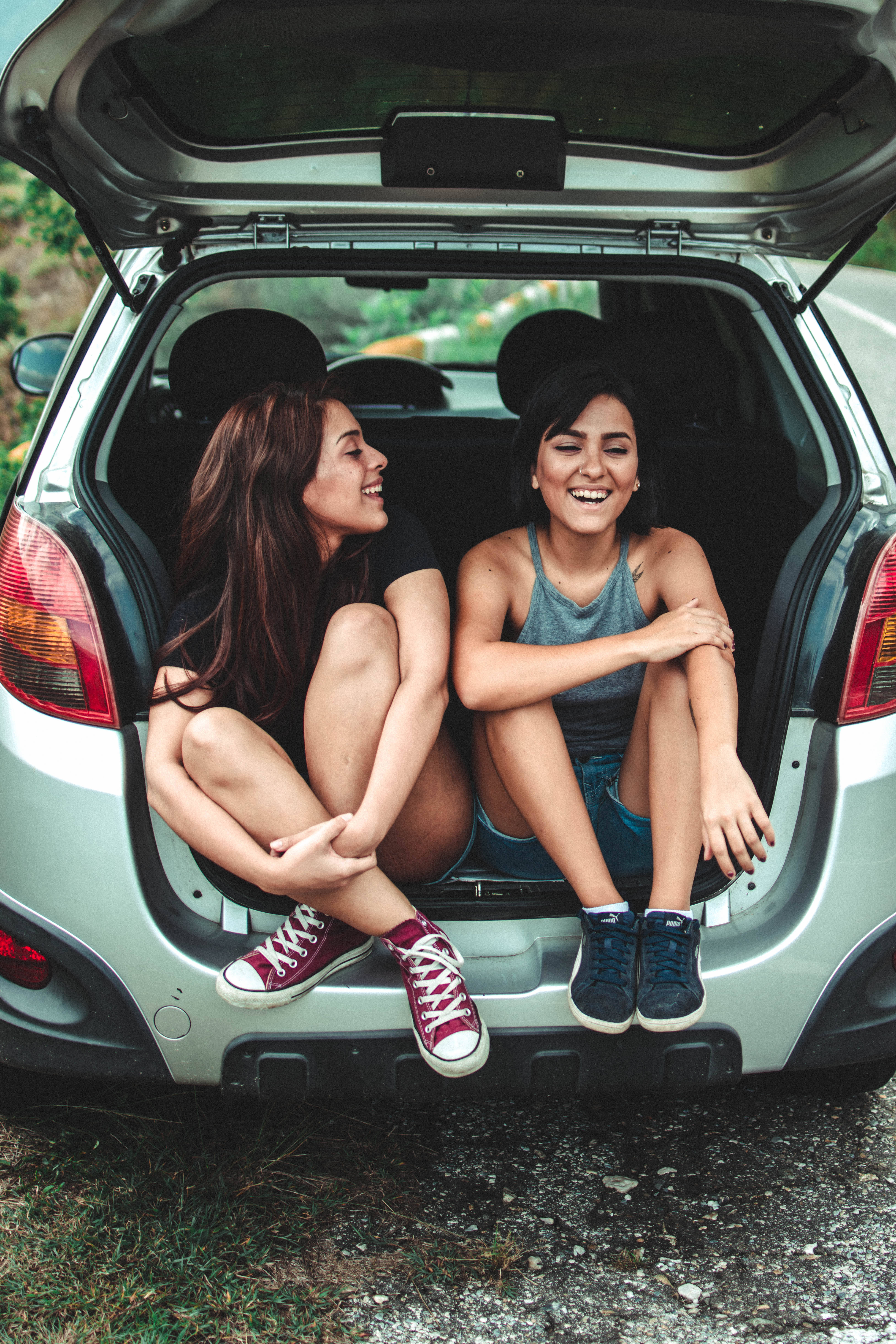 road trip 桌布 壁紙 — 兩位微笑的年輕女性盤腿坐在一輛銀色汽車的打開後車廂中，一起在戶外大笑。