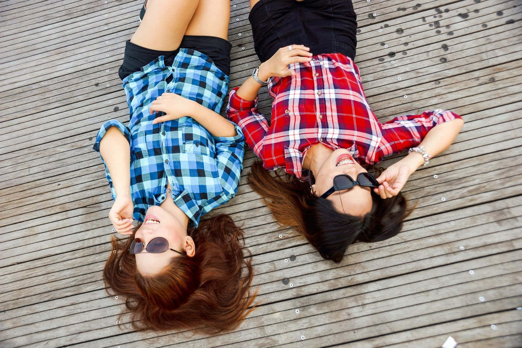 Summer Fashion 桌布 壁紙 — 兩位微笑的年輕女性穿著格紋襯衫，戴著太陽眼鏡，並排躺在木甲板上大笑。