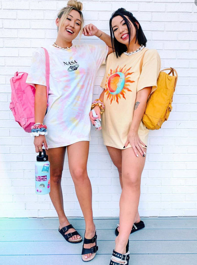 Clothes 桌布 壁紙 — 兩位微笑的年輕女性穿著色彩繽紛的寬鬆T恤和涼鞋，背著鮮豔的背包，在白色磚牆前擺姿勢。