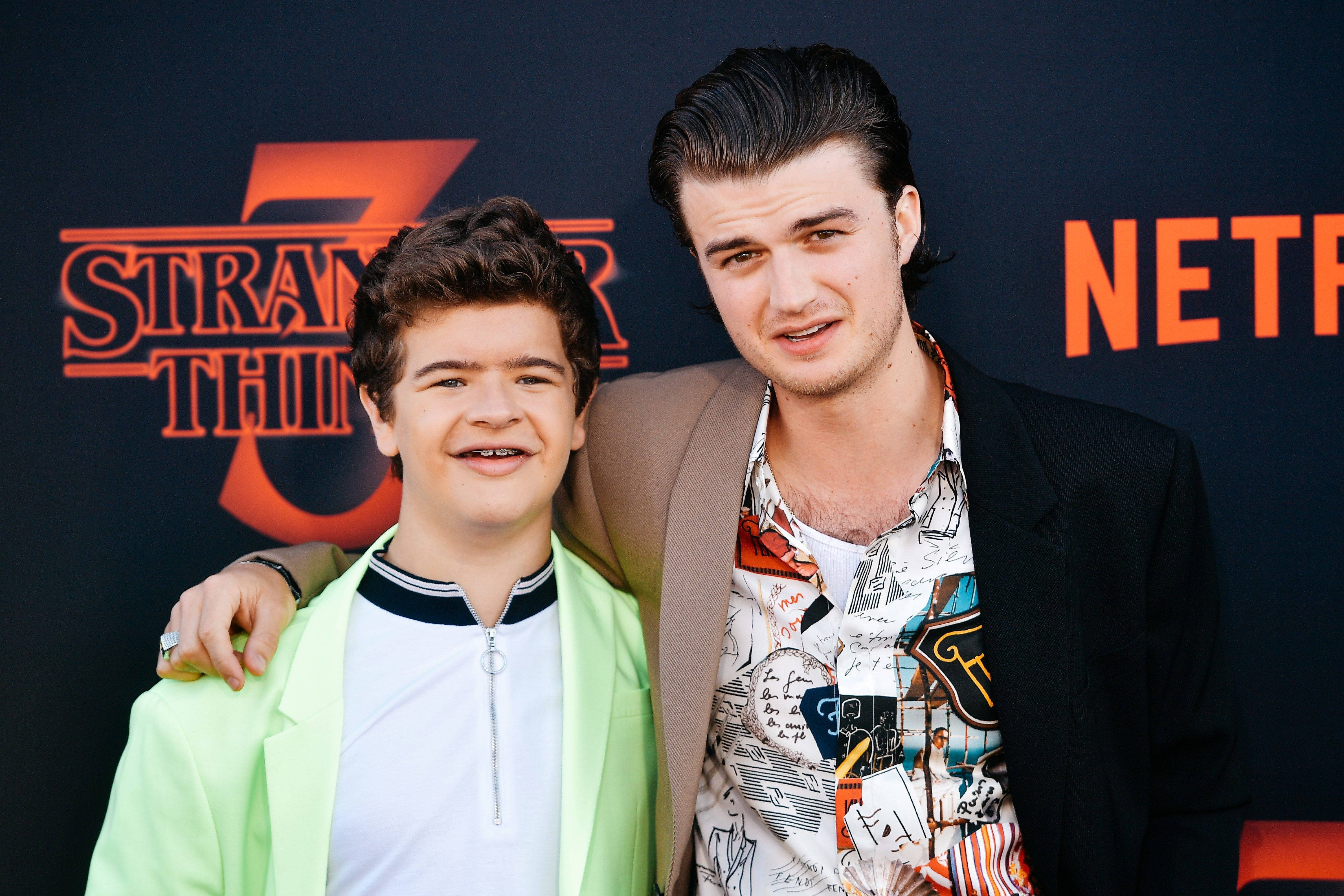 Joe Keery 桌布 壁紙 — 兩位微笑的年輕男子在Stranger Things活動中一起擺姿勢，背景帶有品牌標誌。