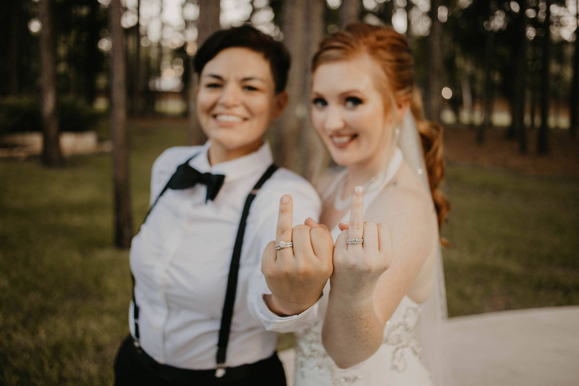 lesbians 桌布 壁紙 — 兩位微笑的新娘穿著婚紗，在陽光明媚的森林中挑釁地向鏡頭展示她們的訂婚戒指。
