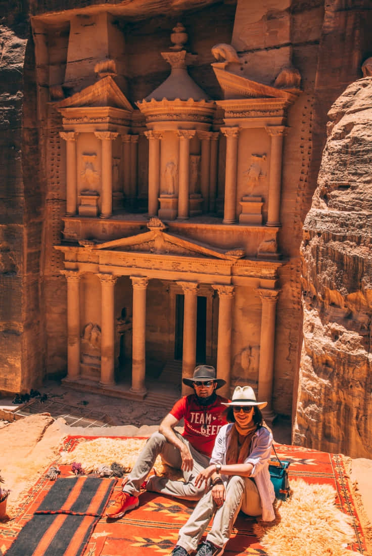 Jordan 桌布 壁紙 — 兩位微笑的旅行者坐在色彩繽紛的地毯上，面前是佩特拉玫瑰色砂岩懸崖中雕刻出的雄偉、陽光照耀的寶庫。