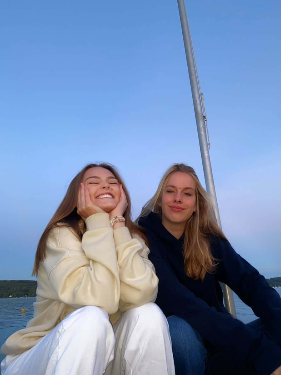 Best Friend Aesthetic 桌布 壁紙 — 兩位微笑的朋友在黃昏時分於帆船上放鬆，身後是平靜的水面和清澈的藍天。