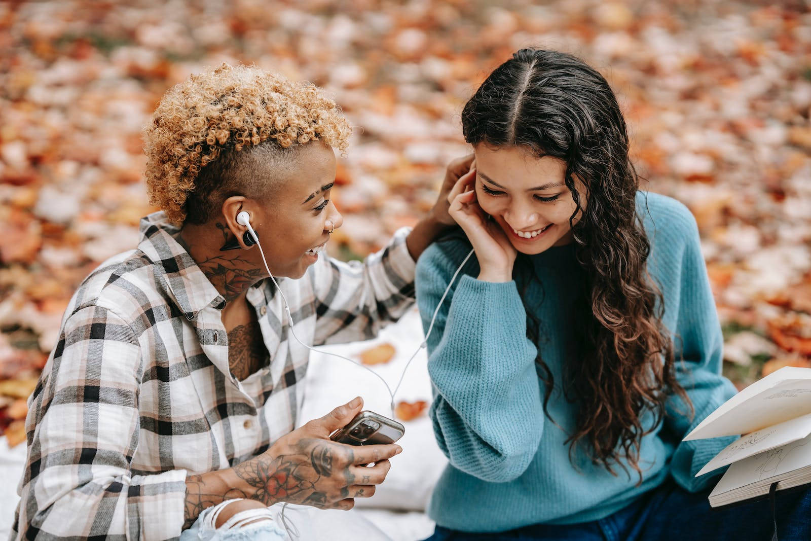 Best Autumn 桌布 壁紙 — 兩位微笑的朋友坐在鋪滿秋葉的地毯上的毯子上，透過耳機分享音樂。