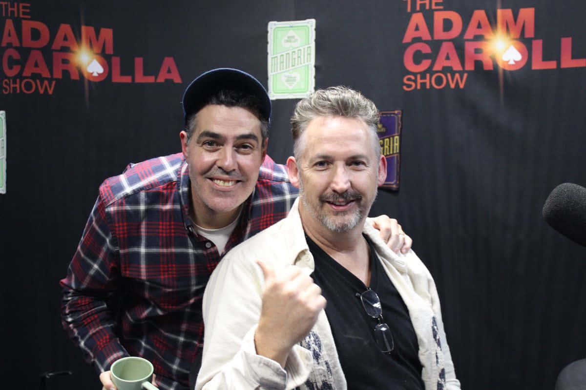  壁紙 — 兩位微笑的男士在 The Adam Carolla Show 中一起擺姿勢，身後有麥克風和品牌背景。