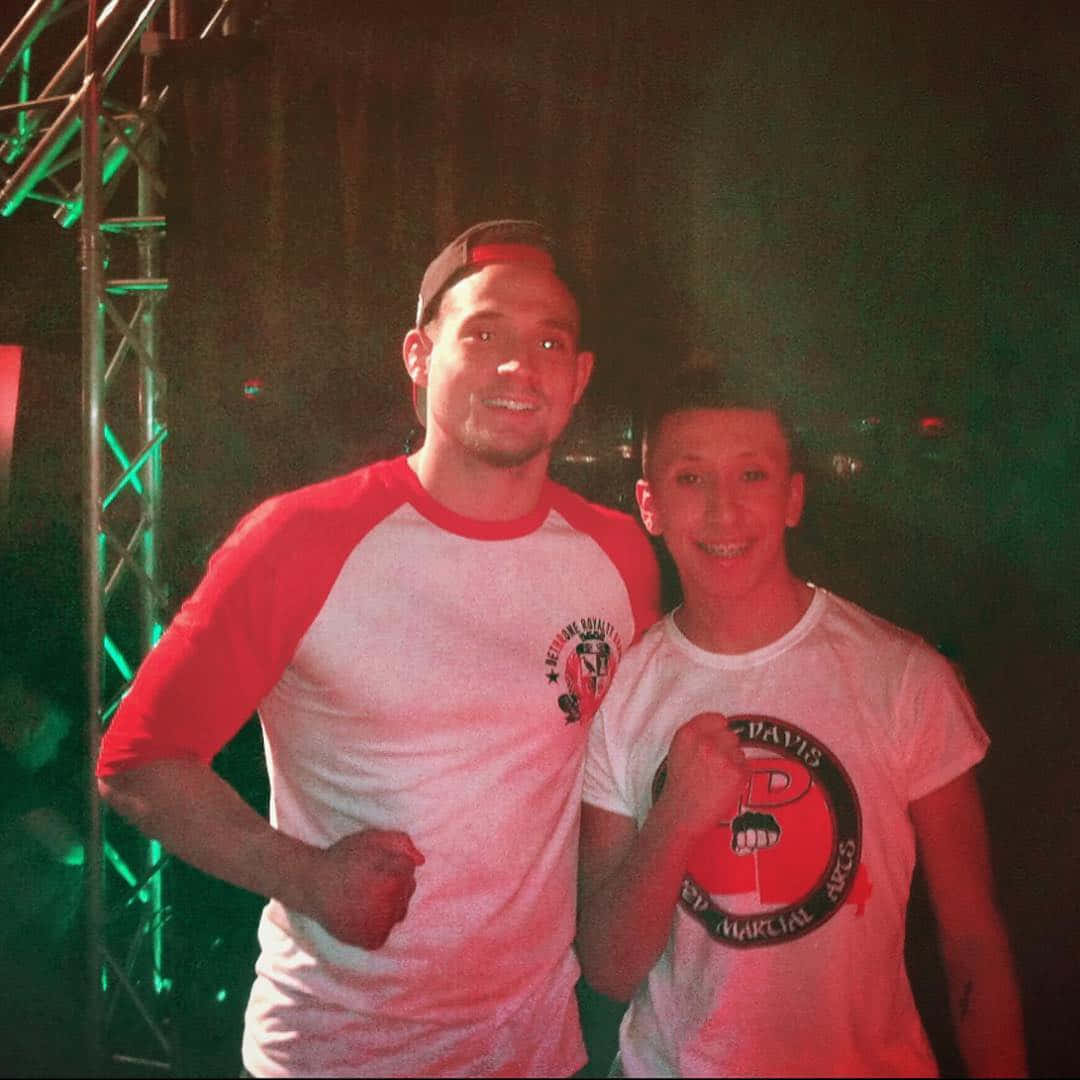 MMA 桌布 壁紙 — 兩位微笑的男士在燈光昏暗的活動中一起擺姿勢，背景有綠色舞台燈光和金屬桁架結構。