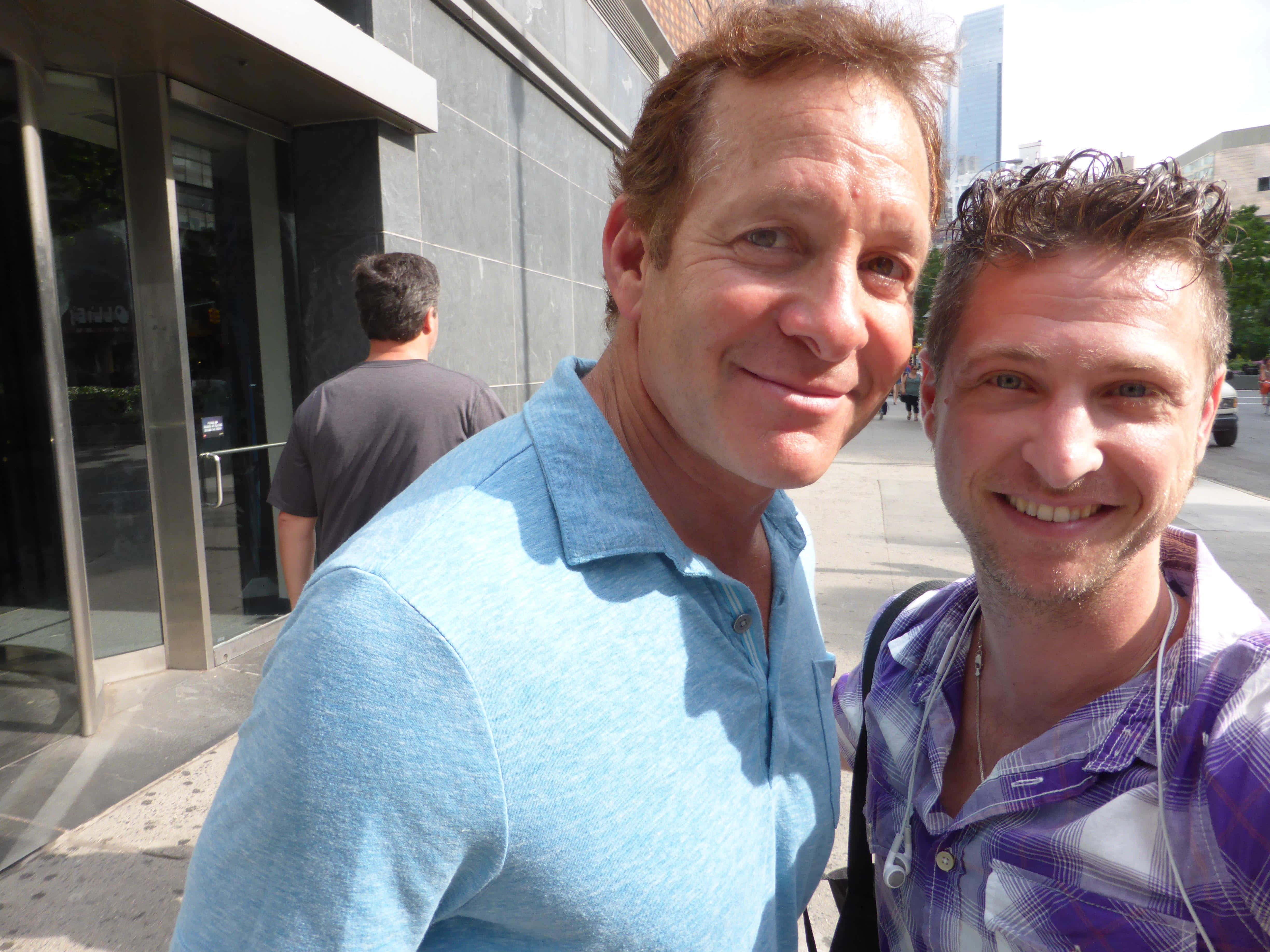 Steve Guttenberg 桌布 壁紙 — 兩位微笑的男士在陽光明媚的城市人行道上，在現代玻璃建築入口附近自拍。