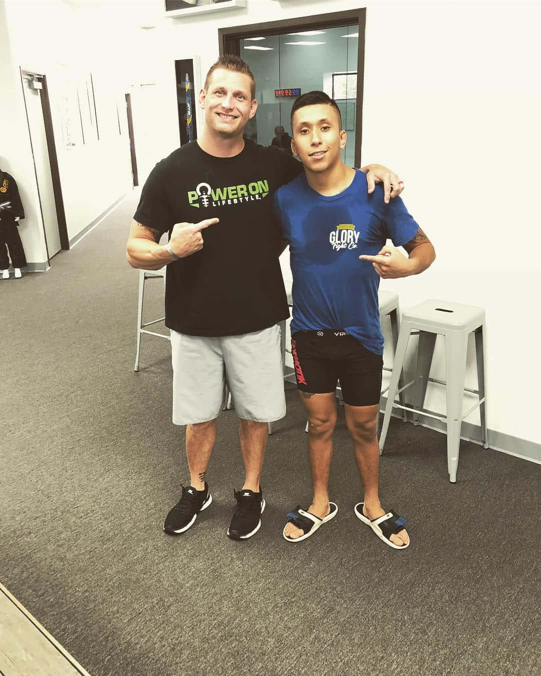 健身房 桌布 壁紙 — 兩位微笑的男士穿著健身服裝在走廊一起擺姿勢，指著他們的品牌襯衫。