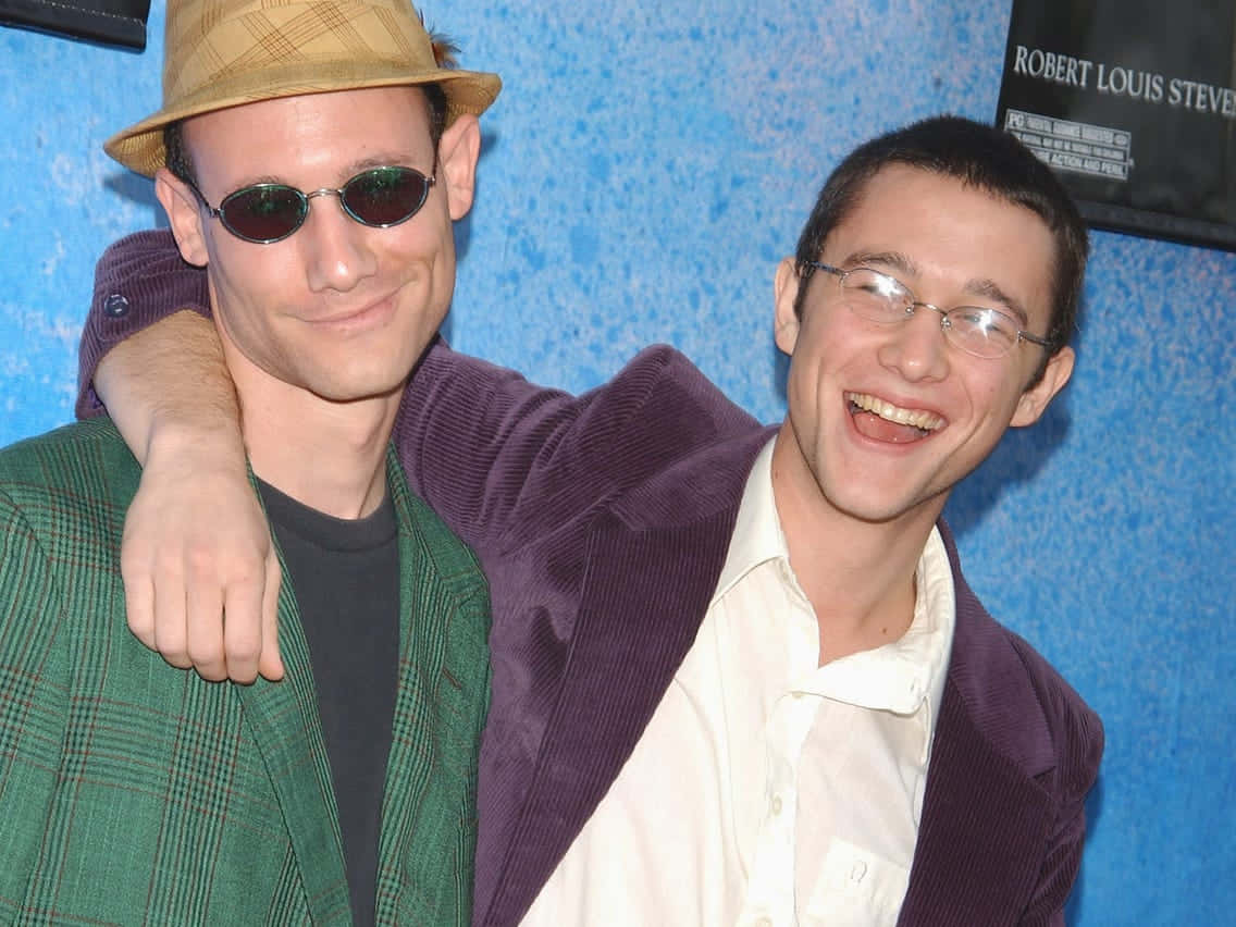 joseph gordon-levitt 桌布 壁紙 — 兩位微笑的男子一起擺姿勢，一位身穿綠色格紋外套戴帽，另一位身穿紫色西裝外套，背景是藍色紋理牆。