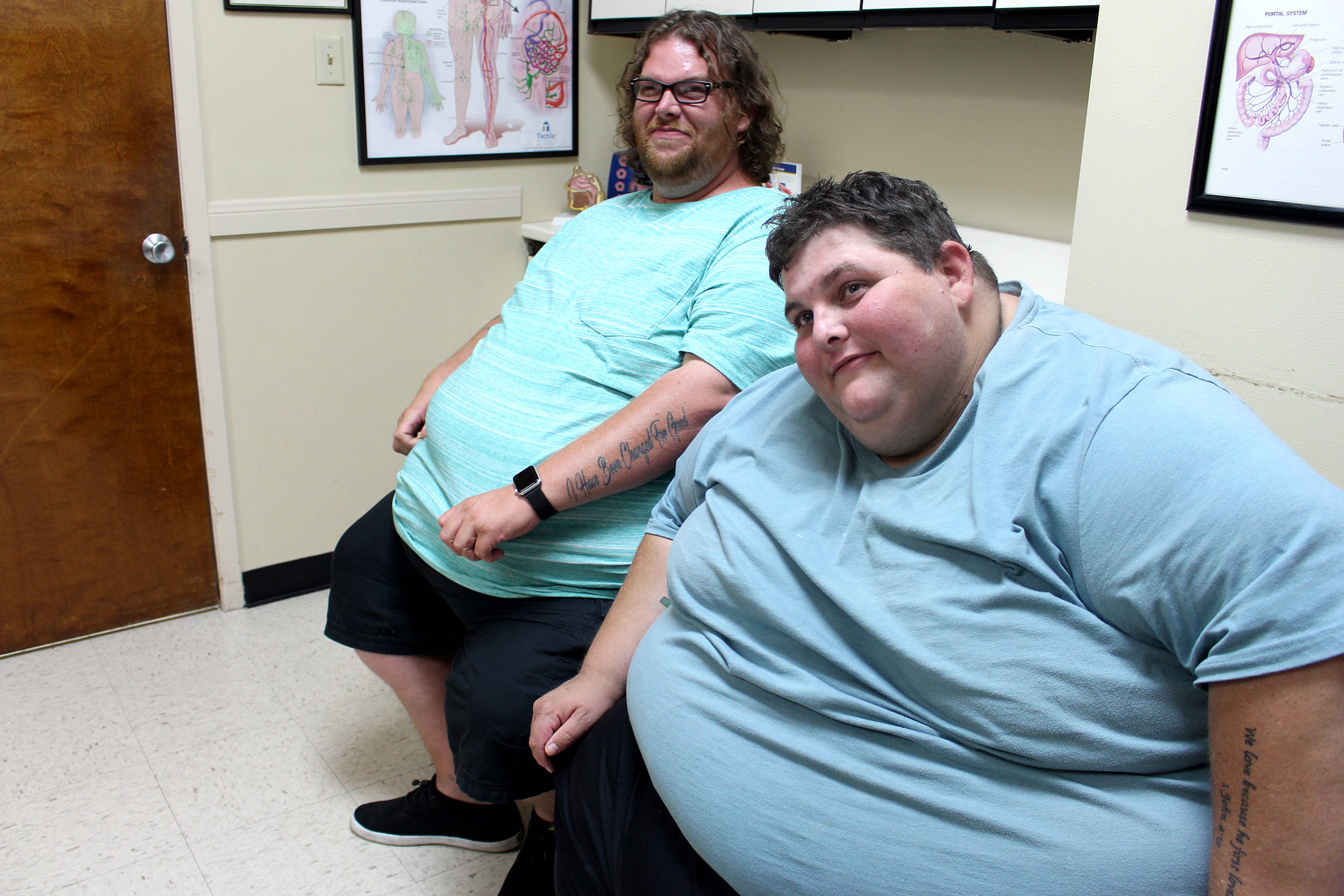 fat 桌布 壁紙 — 兩位微笑的男子身穿淺藍色襯衫，並排坐在一間牆上掛有解剖學海報的醫療檢查室中。