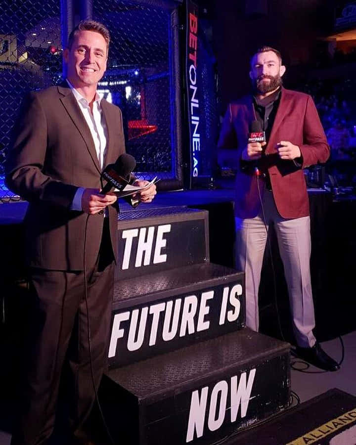 UFC 桌布 壁紙 — 兩位微笑的男評論員身穿西裝，站在樓梯旁，樓梯上有粗體白色文字寫著「THE FUTURE IS NOW」，在藍色舞台燈光下的UFC賽事中。