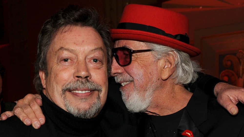 Tim Curry 桌布 壁紙 — 兩位微笑的白鬍子男士，其中一位戴著紅帽子和太陽眼鏡，在昏暗的室內環境中熱情擁抱。