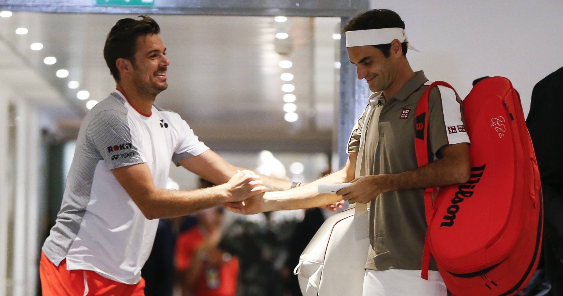 Tennis 桌布 壁紙 — 兩位微笑的網球運動員在光線明亮的室內走廊握手，其中一人背著紅色Wilson球拍袋。
