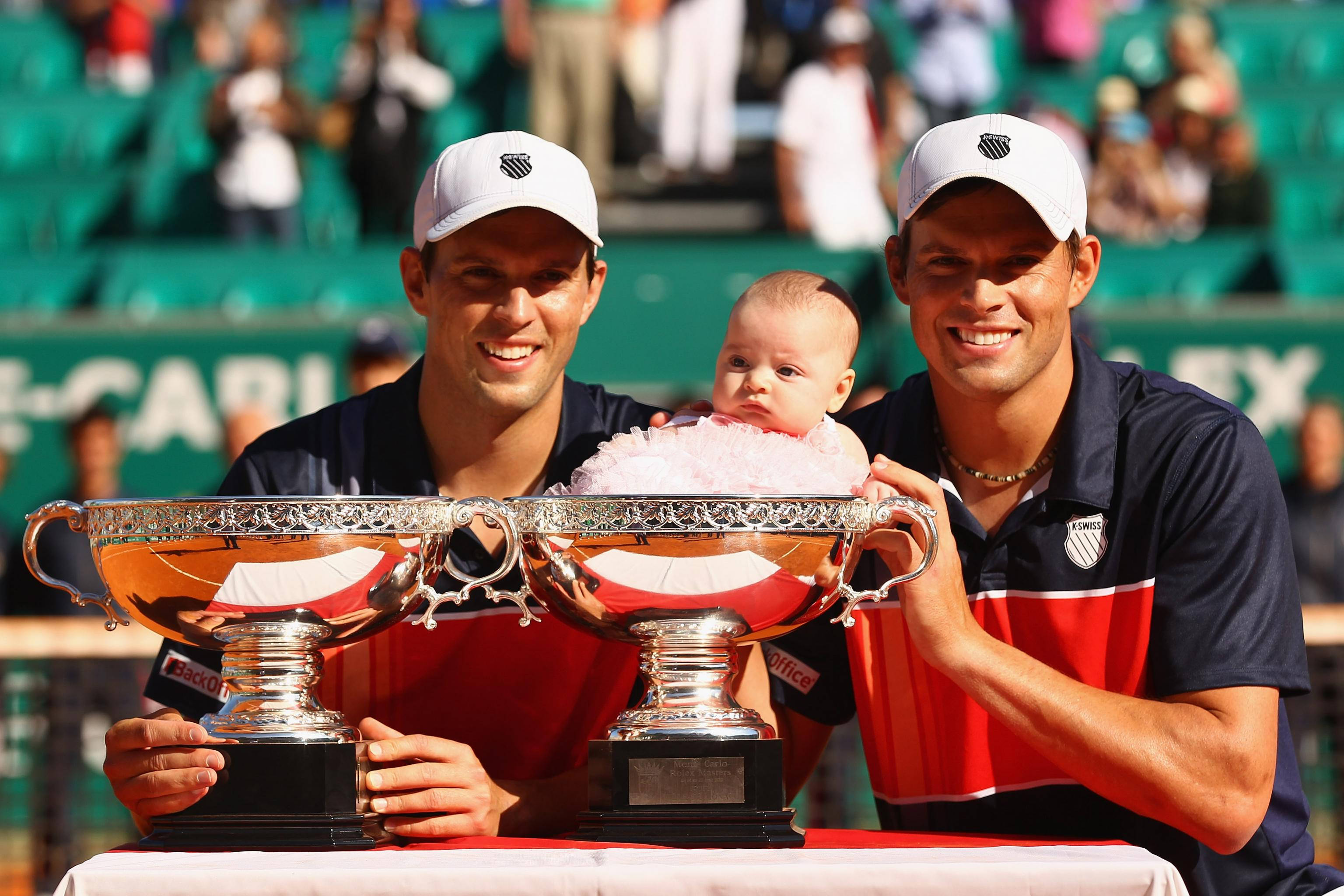 Tennis 桌布 壁紙 — 兩位微笑的網球選手手持大型銀色獎盃，同時在獎盃中央懷抱著一個嬰兒。