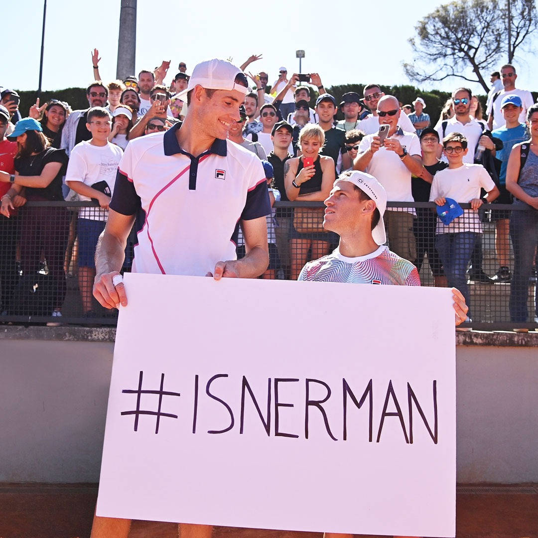 網球 桌布 壁紙 — 兩位微笑的網球選手舉著 #ISNERMAN 標語牌，粉絲們在圍欄後歡呼，陽光燦爛。