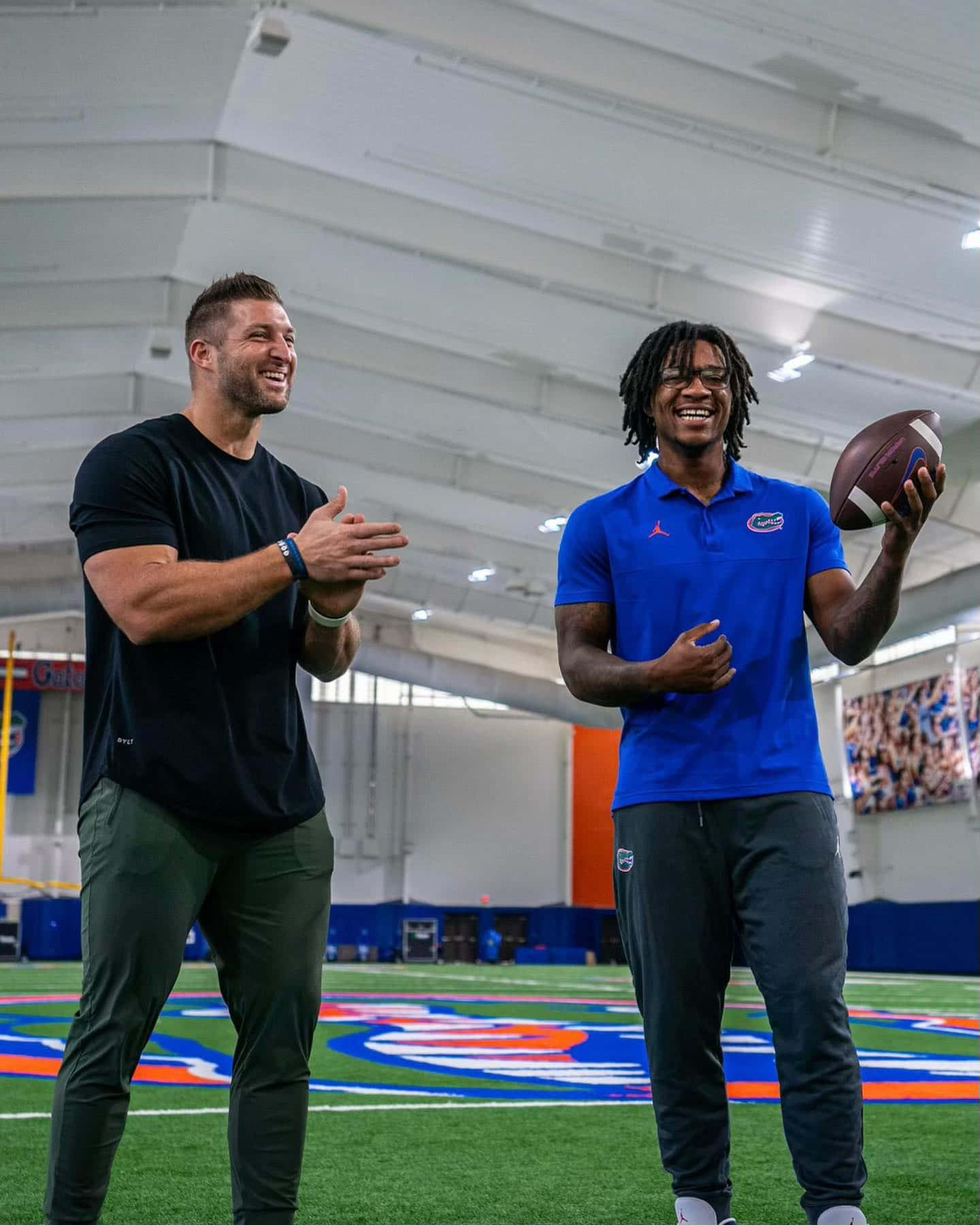 Tim Tebow 桌布 壁紙 — 兩位微笑的足球運動員在室內分享歡樂時光，一人拿著足球，另一人在充滿活力的草地球場上鼓掌。