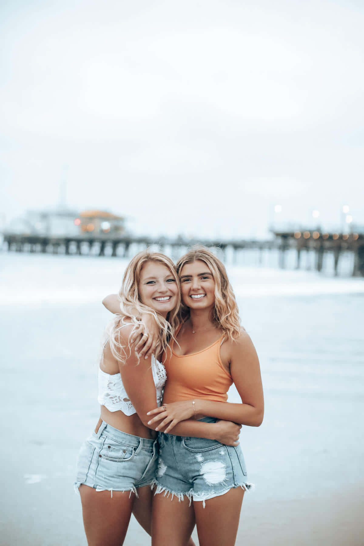 Friends 桌布 壁紙 — 兩位微笑的金髮女性在沙灘上擁抱，背景有碼頭和柔和天空。