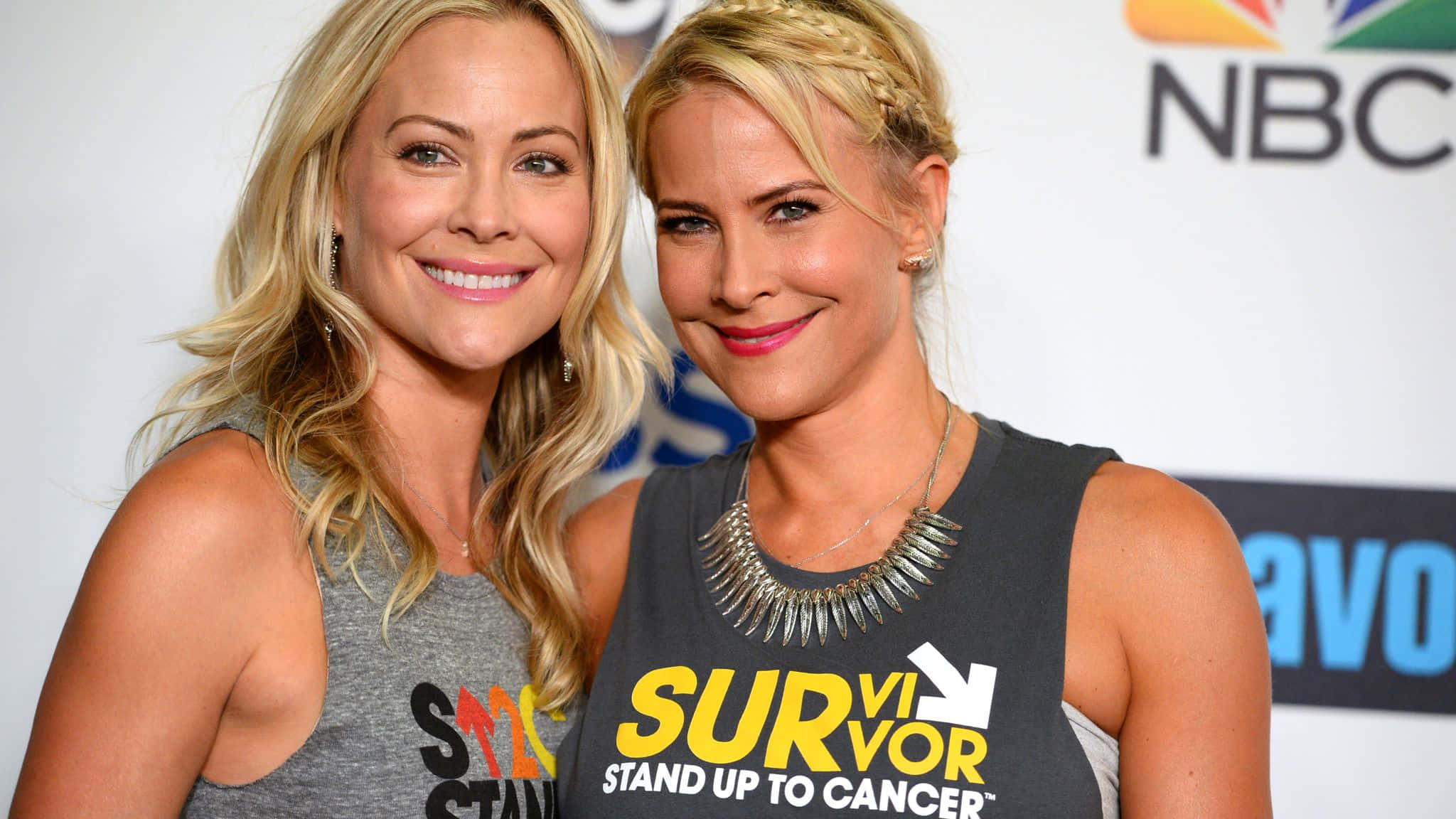 Brittany Daniel 桌布 壁紙 — 兩位微笑的金髮女性在癌症意識活動中穿著品牌T恤，在白色背景前與NBC標誌一起合影。