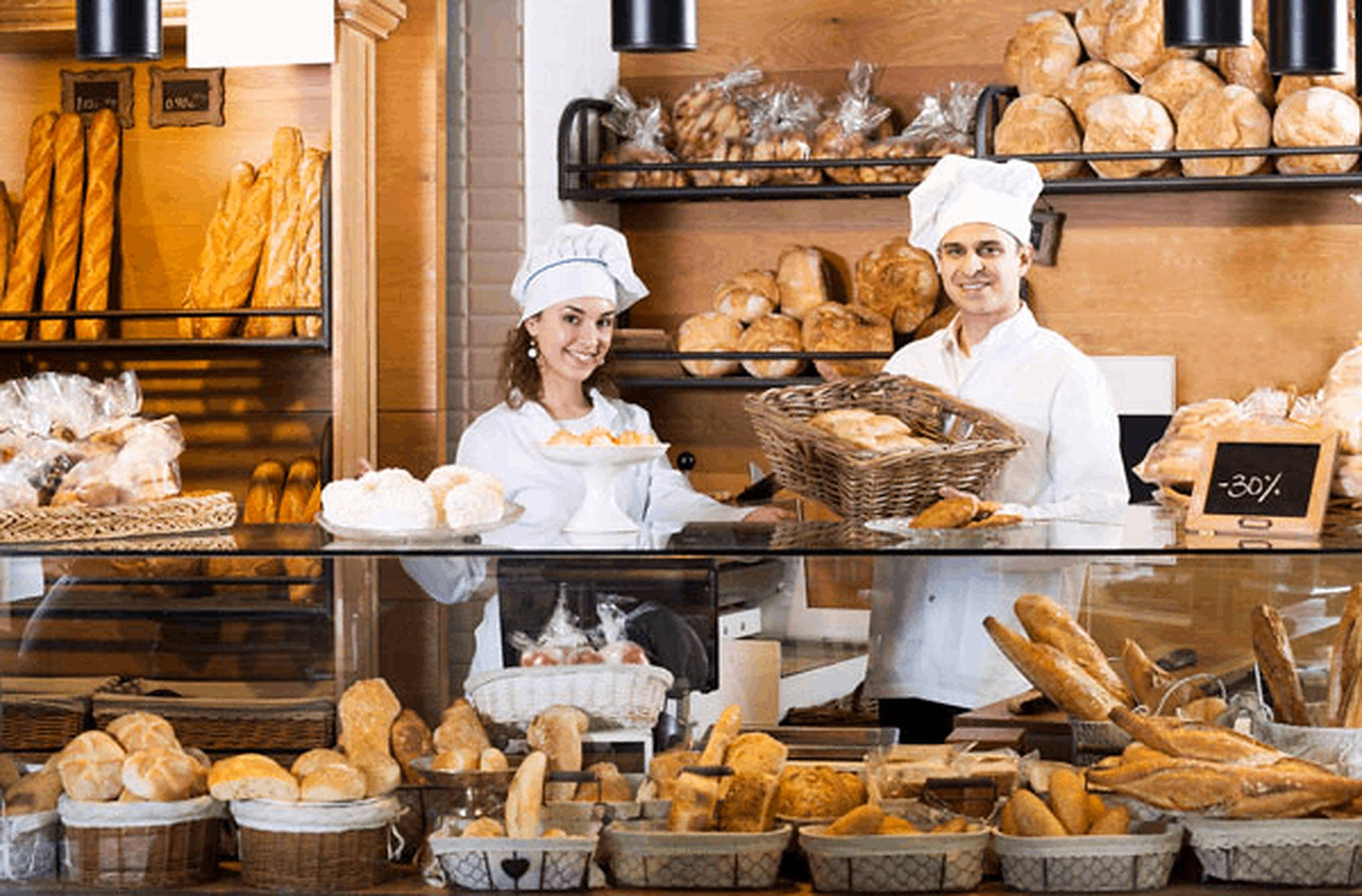 Bakery 桌布 壁紙 — 兩位微笑的麵包師身穿白色制服，在玻璃麵包櫃後自豪地展示新鮮金黃色的麵包。