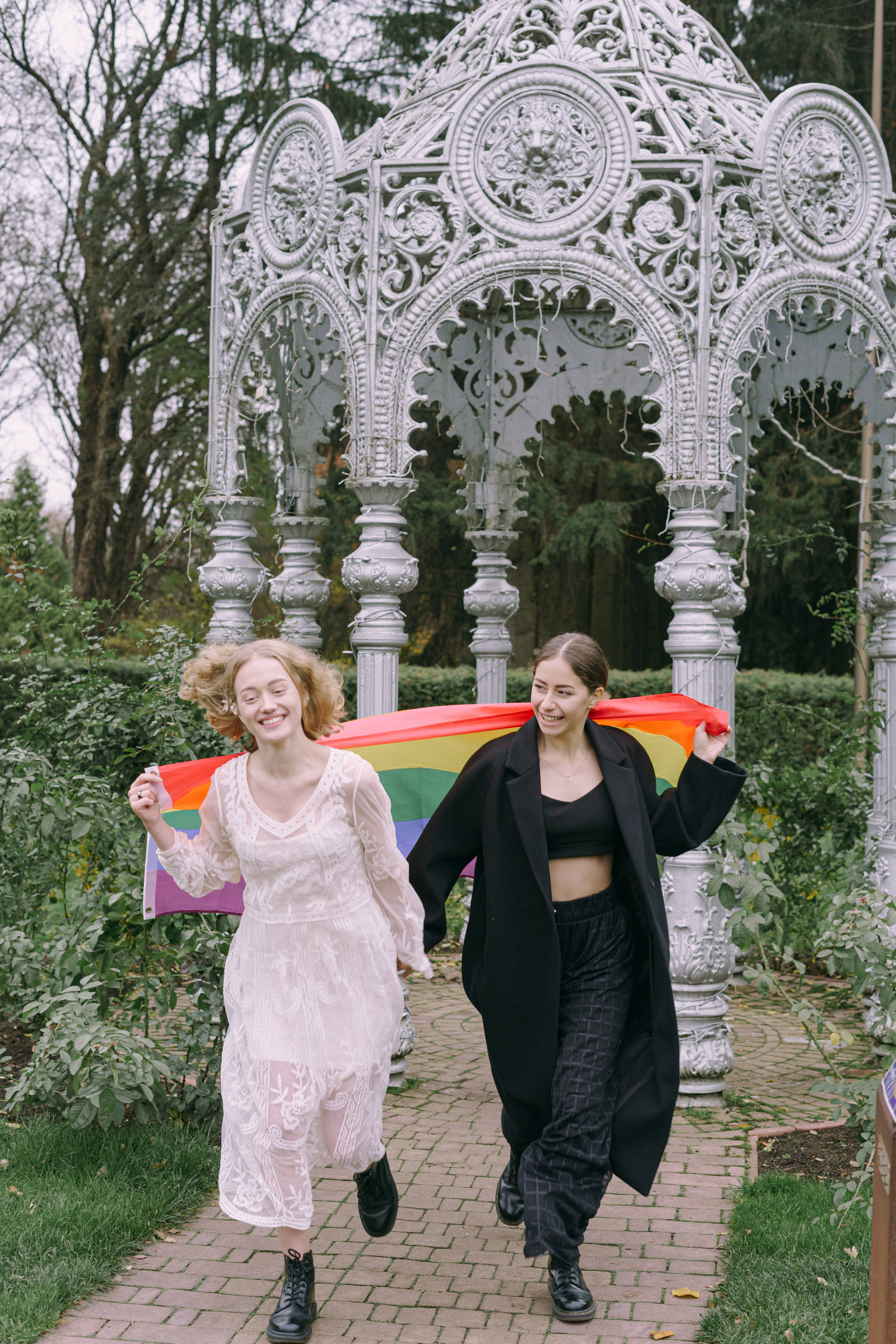 Pride Flag 桌布 壁紙 — 兩位快樂的女性在裝飾華麗的白色涼亭下，手持彩虹旗跑過花園。