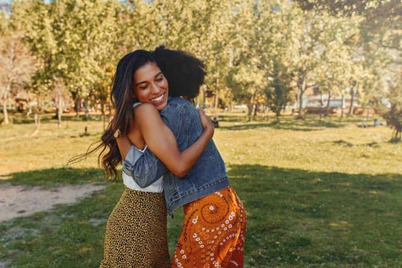 Hug 桌布 壁紙 — 兩位快樂的女性在陽光明媚的公園裡熱情擁抱，身後是金色的樹木和綠色的草地。