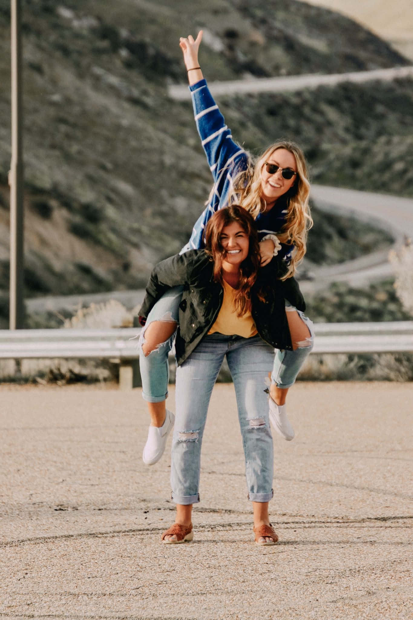 Friends 桌布 壁紙 — 兩位快樂的女性在陽光明媚的山路上分享一個有趣的揹背時刻，身後是蜿蜒的瀝青路和岩石丘陵。