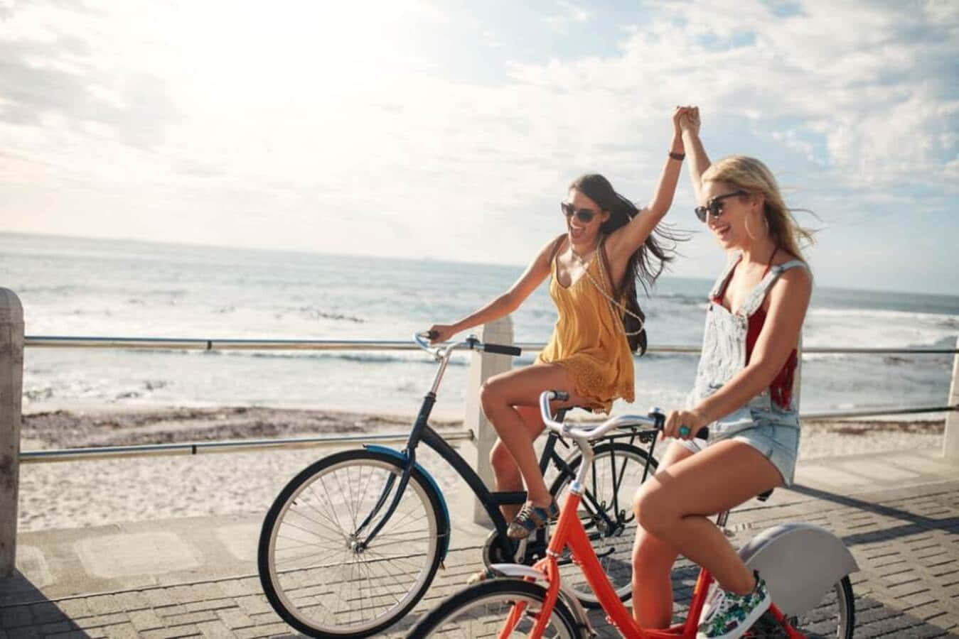 Beach Cruiser 桌布 壁紙 — 兩位快樂的女性沿著海邊木板路騎自行車，身後的海浪拍打著。