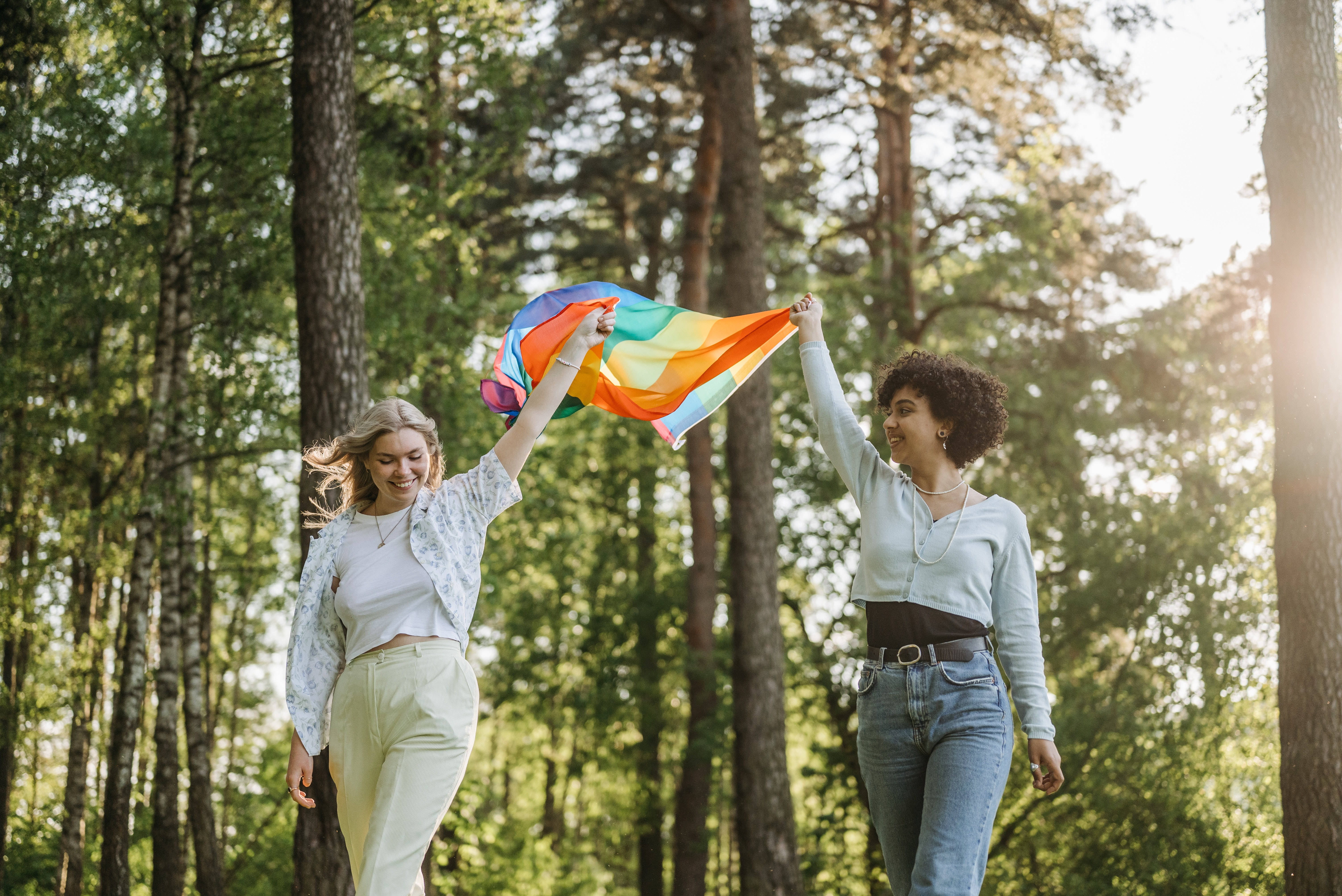 女同志旗幟 桌布 壁紙 — 兩位快樂的女性穿過陽光普照的森林，高舉著鮮豔的彩虹旗。