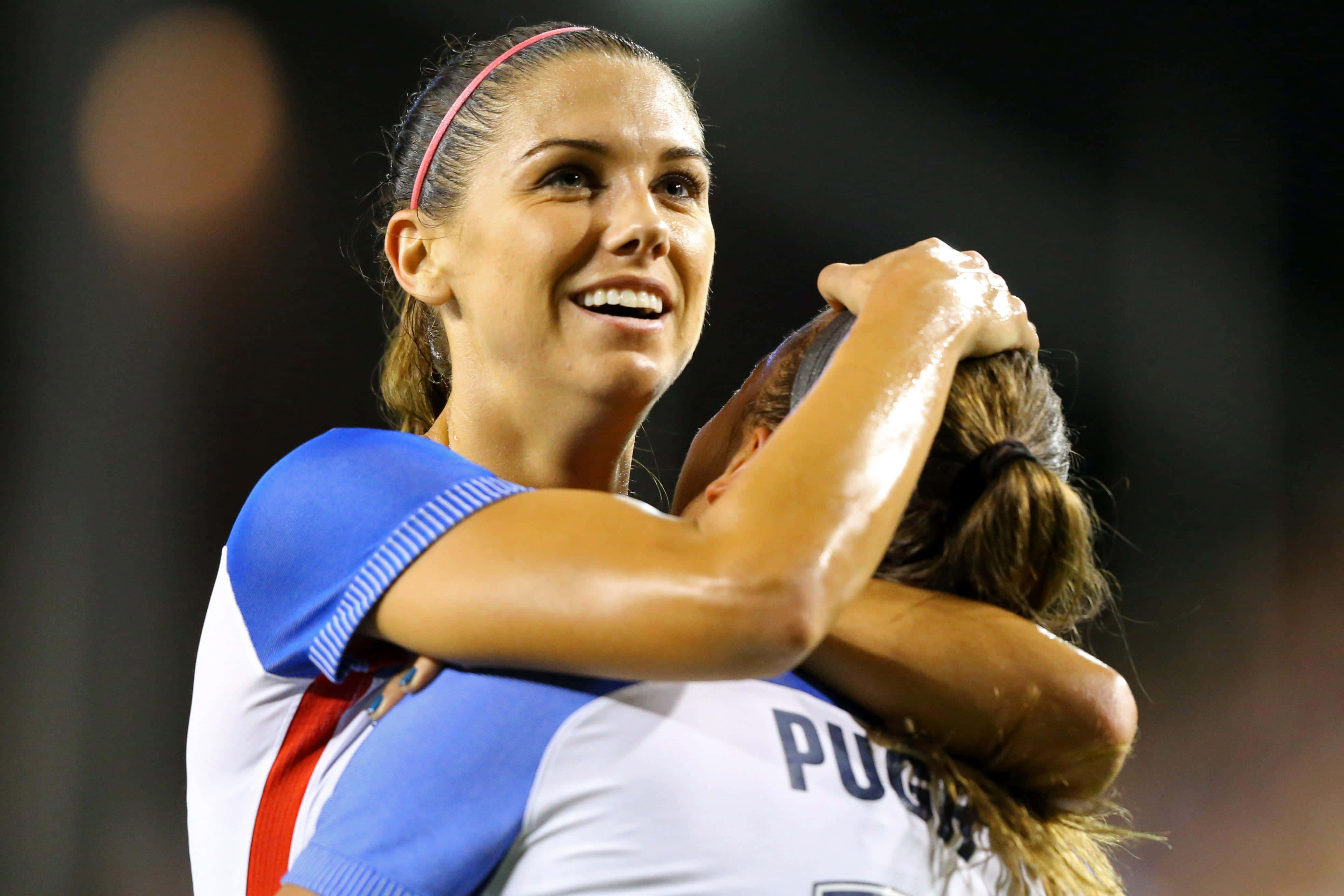 Alex Morgan 桌布 壁紙 — 兩位快樂的女足球員在明亮的燈光下於體育場場地上勝利後緊緊擁抱。