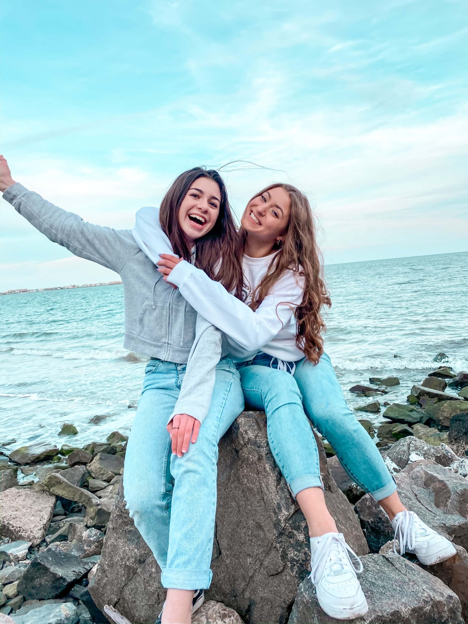 Friends 桌布 壁紙 — 兩位快樂的年輕女性在柔和藍天下，於岩石海邊懸崖上一起大笑並擺姿勢。
