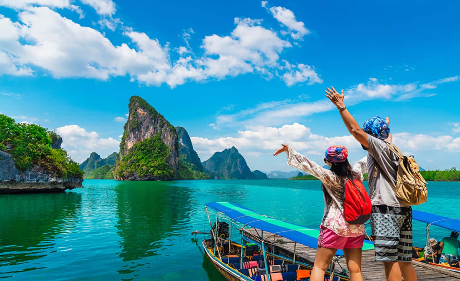 泰國 桌布 壁紙 — 兩位快樂的旅客在長尾船上舉起雙臂，驚嘆於碧綠的海水、高聳的叢林覆蓋的石灰岩喀斯特地貌，以及明亮的藍天。