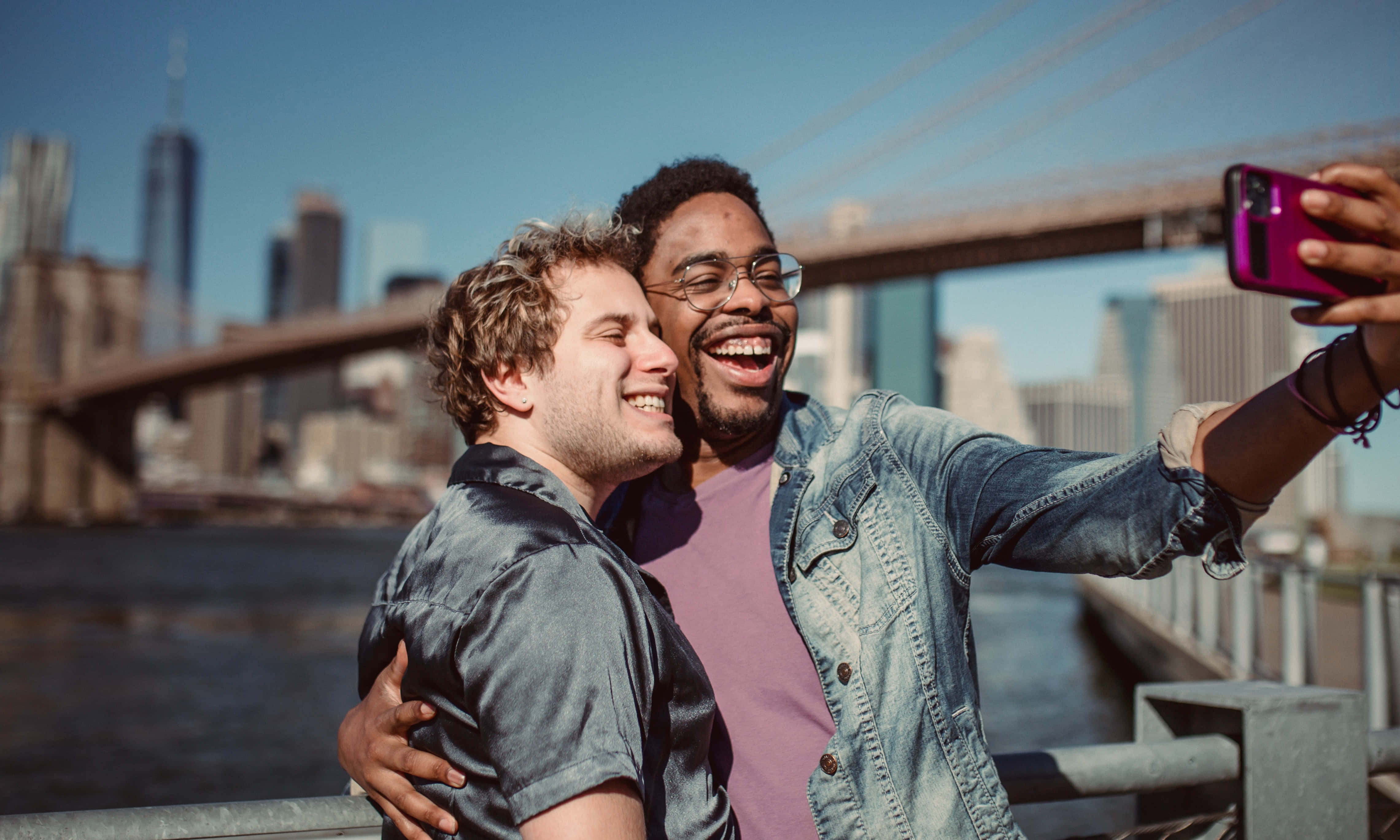 Friendship 桌布 壁紙 — 兩位快樂的朋友在橋上笑著自拍，背景是城市天際線和明亮的藍天。